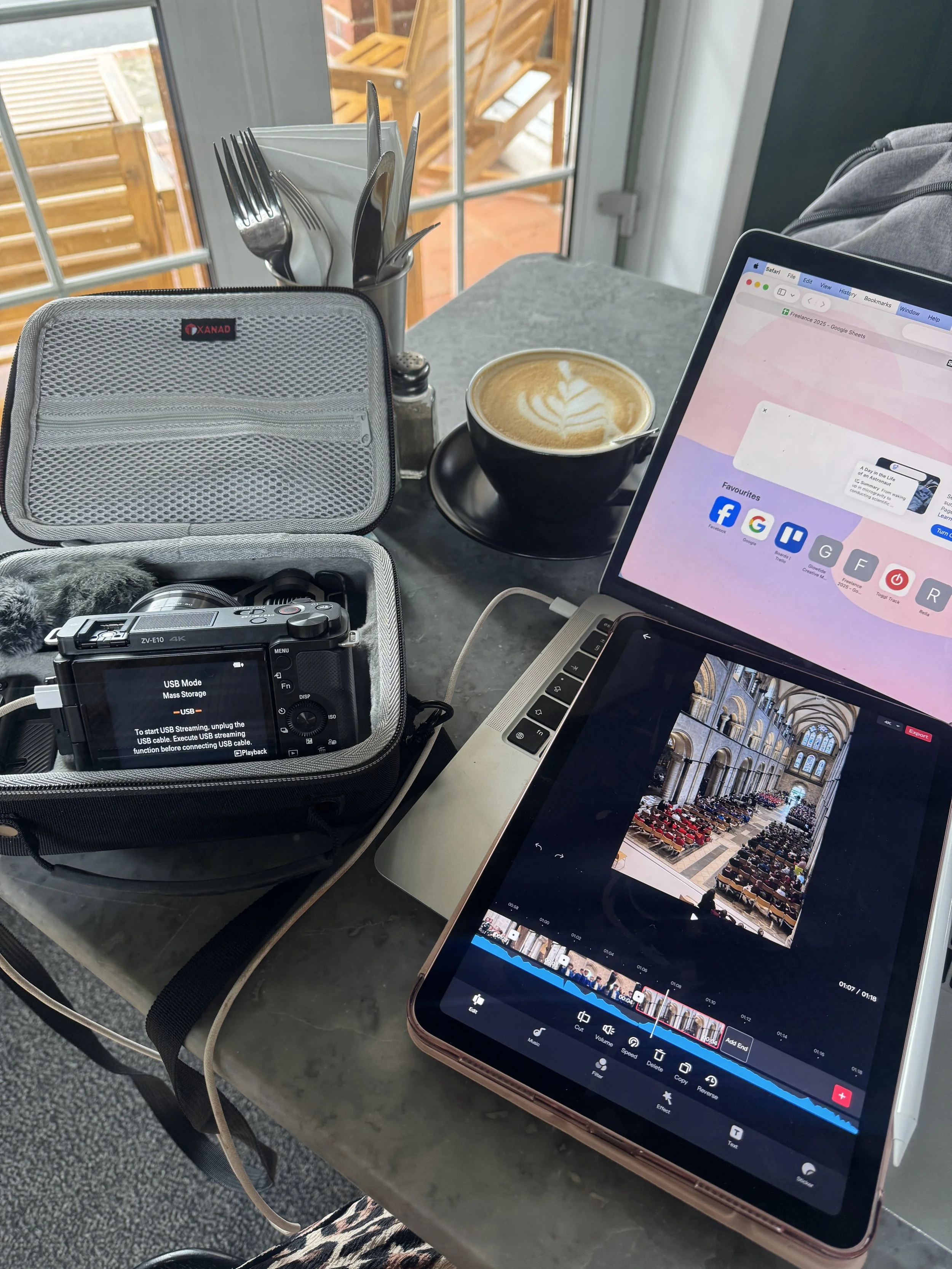 A workspace with a laptop, a tablet, and a camera inside a gray case on a table. There is a cup of coffee with latte art, a container with utensils, and a window in the background showing an outdoor wooden deck.