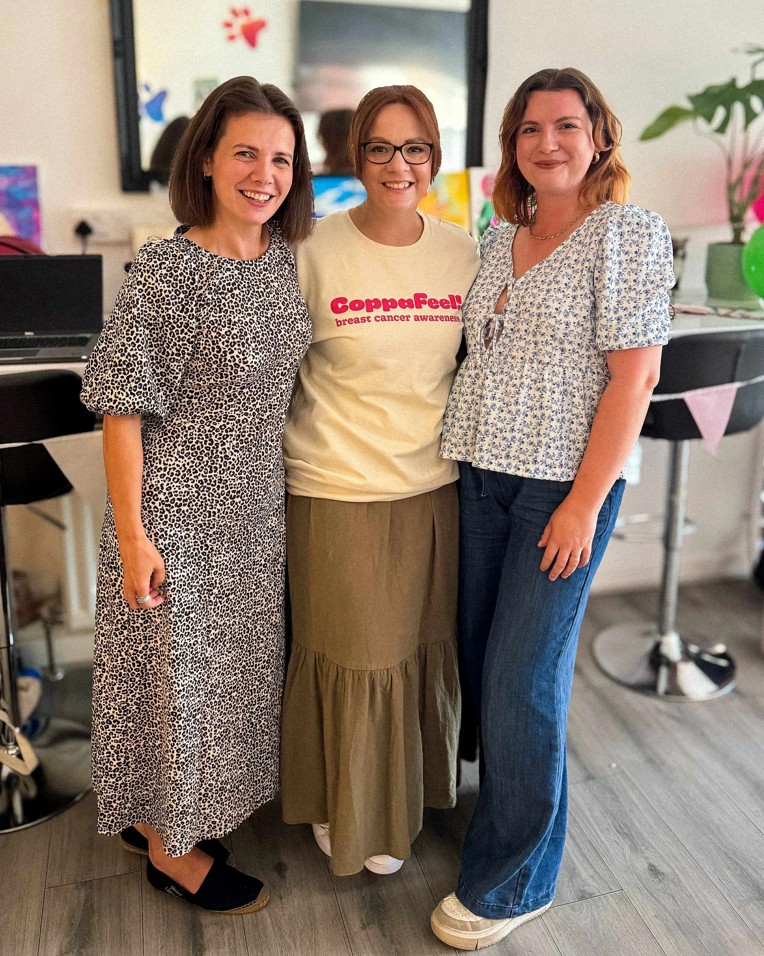Three women standing together indoors, smiling for the camera. The woman on the left is wearing a leopard-print dress, the woman in the middle is wearing glasses and a cream-colored T-shirt with red text that reads 'Coppafeel! breast cancer awareness,' and the woman on the right is wearing a floral blouse and jeans. There are colorful decorations and a mirror in the background.