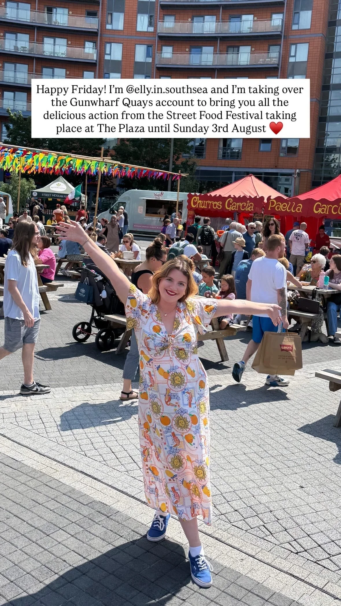 A smiling woman in a colorful, patterned dress with sneakers, arms outstretched, standing in an outdoor food festival with people, food tents, and high-rise buildings in the background.