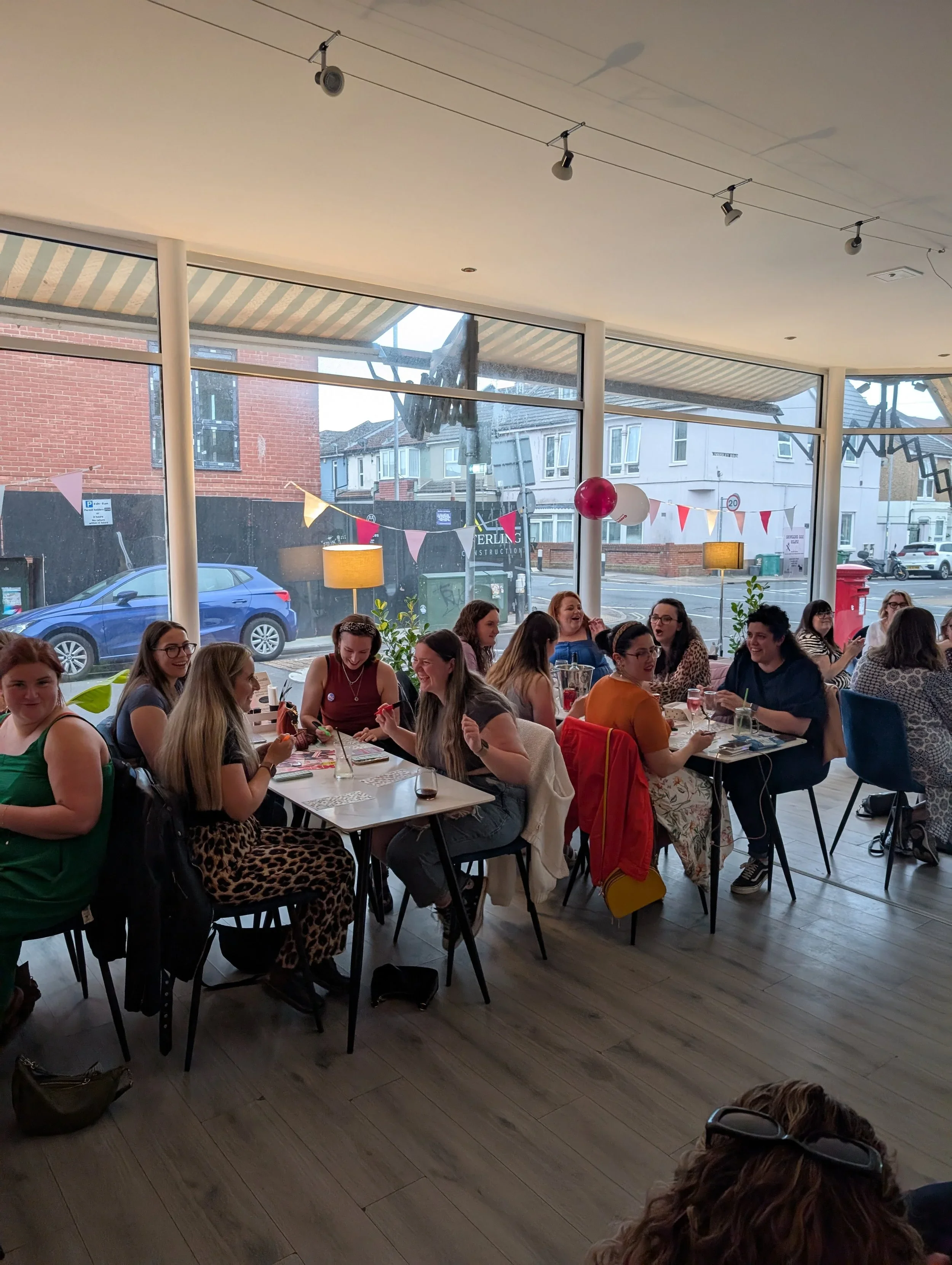 A group of people sitting at tables inside a well-lit cafe or restaurant with large windows showing a street scene outside. The room has simple decor with balloons and bunting hanging from the ceiling.