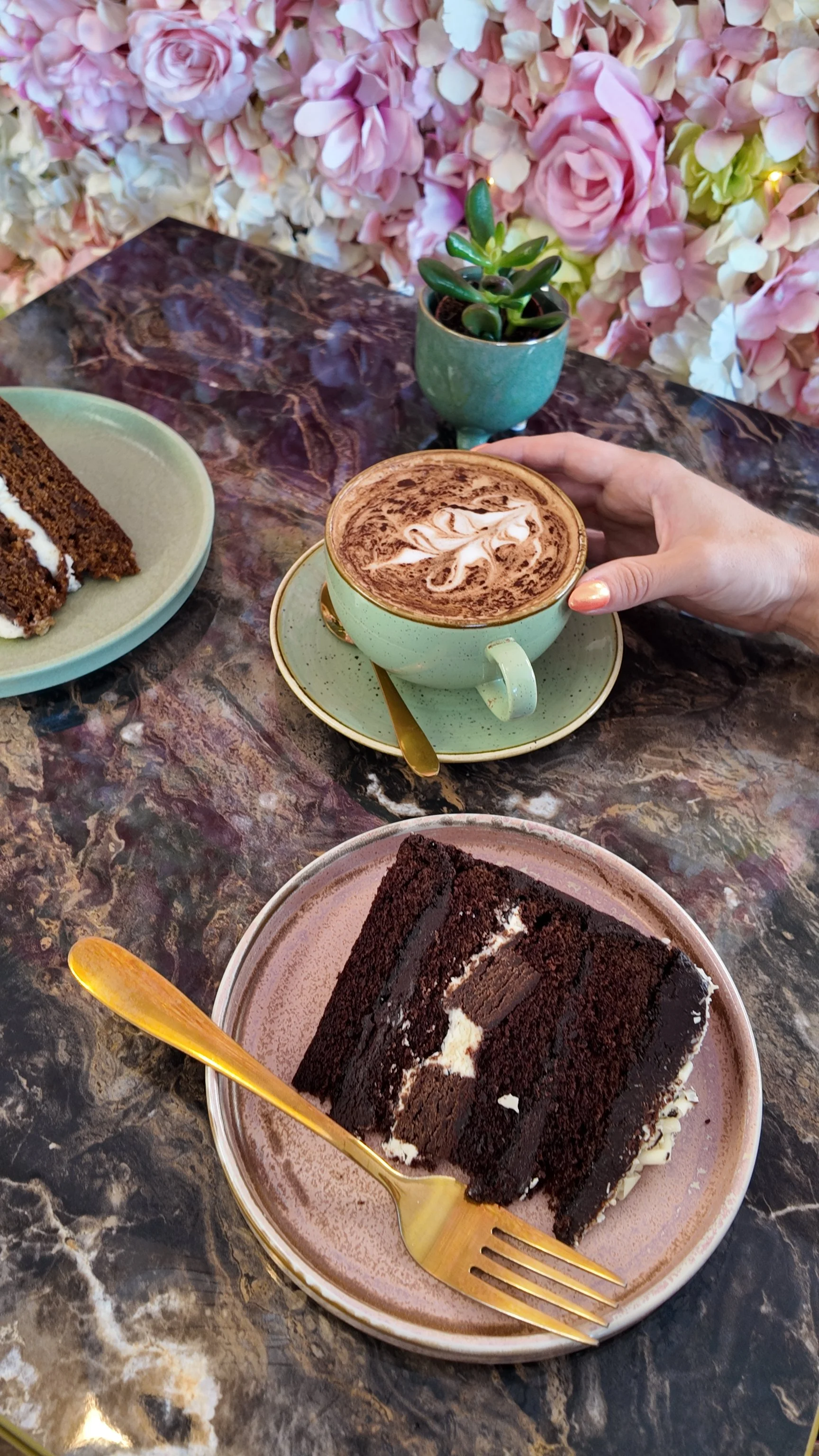 A marble table with slices of chocolate cake, a cup of hot chocolate with whipped cream, a small potted succulent plant, and a floral backdrop.