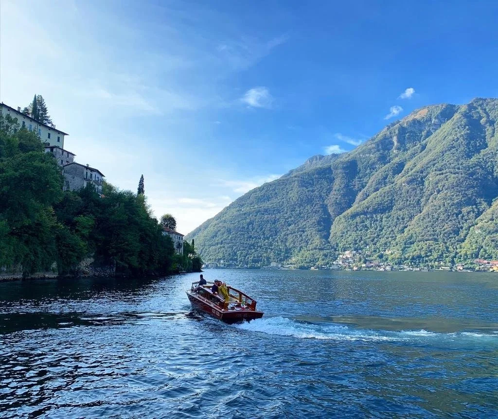 A boat with two people on a lake surrounded by green mountains and trees, under a blue sky.