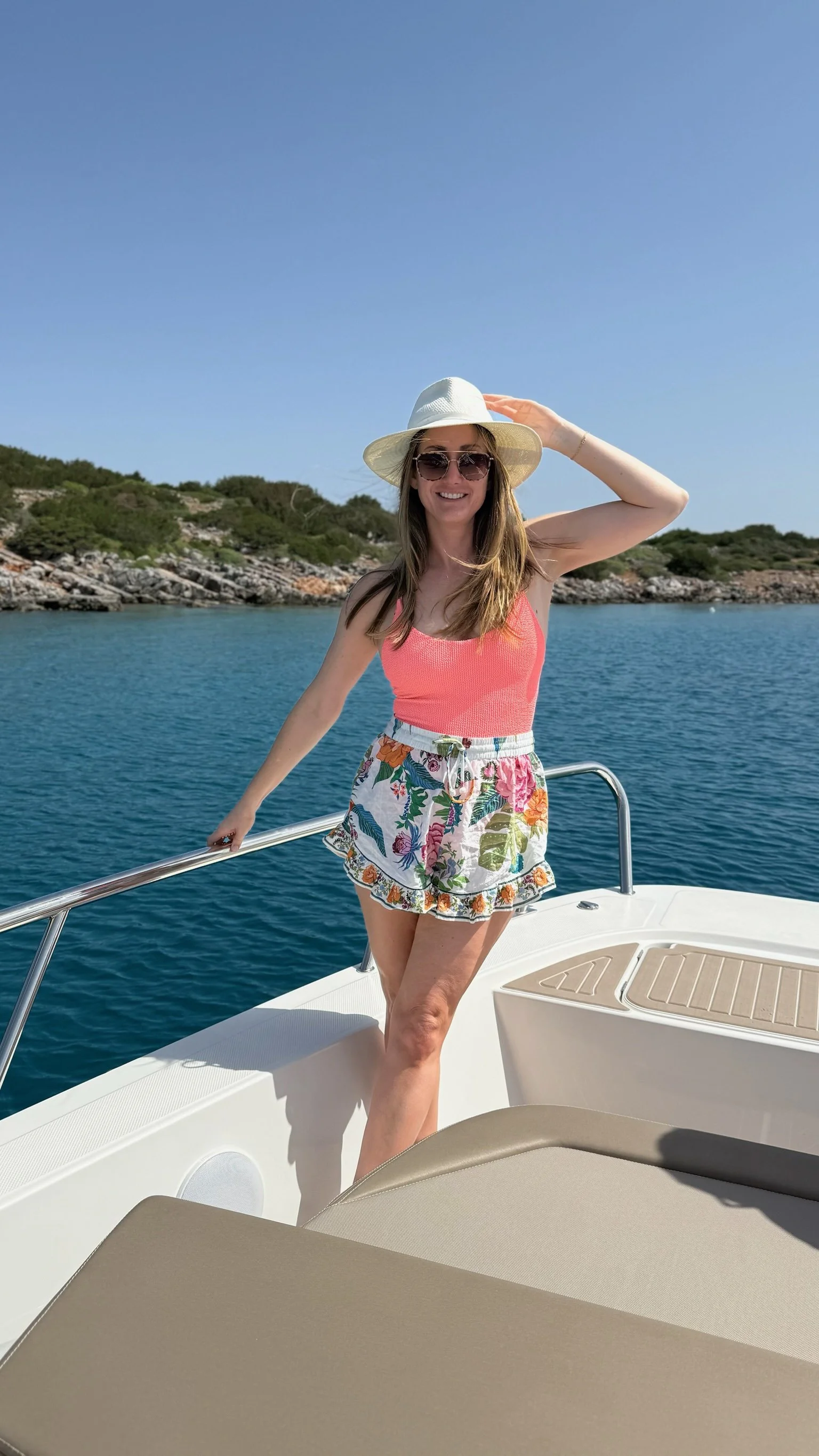 A woman in a coral tank top, floral shorts, a white wide-brim hat, and sunglasses standing on a boat, holding the boat's railing with water and shoreline in the background.