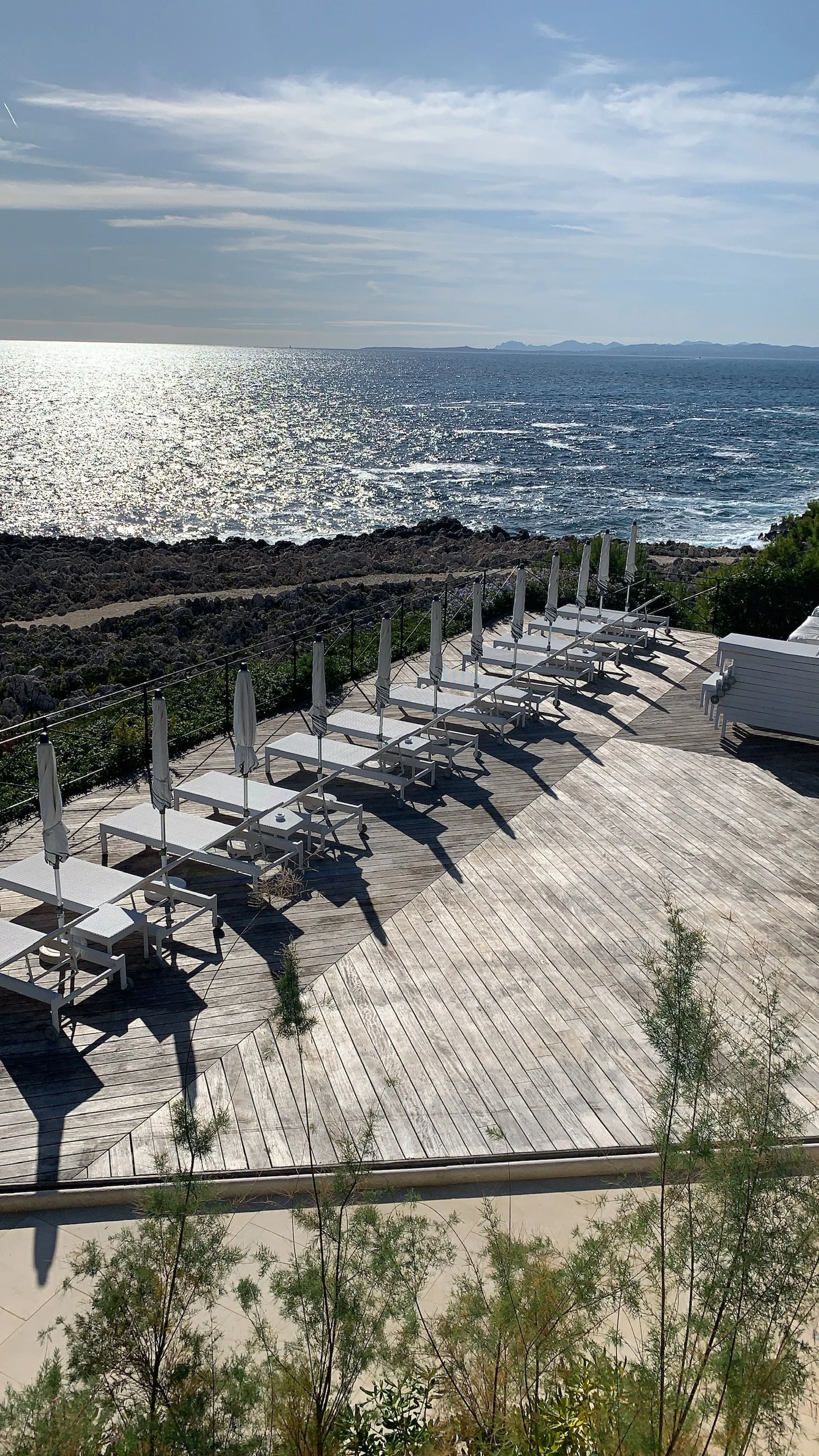 A seaside deck with white lounge chairs and umbrellas overlooking the ocean, with rocky shoreline and distant mountains in the background.