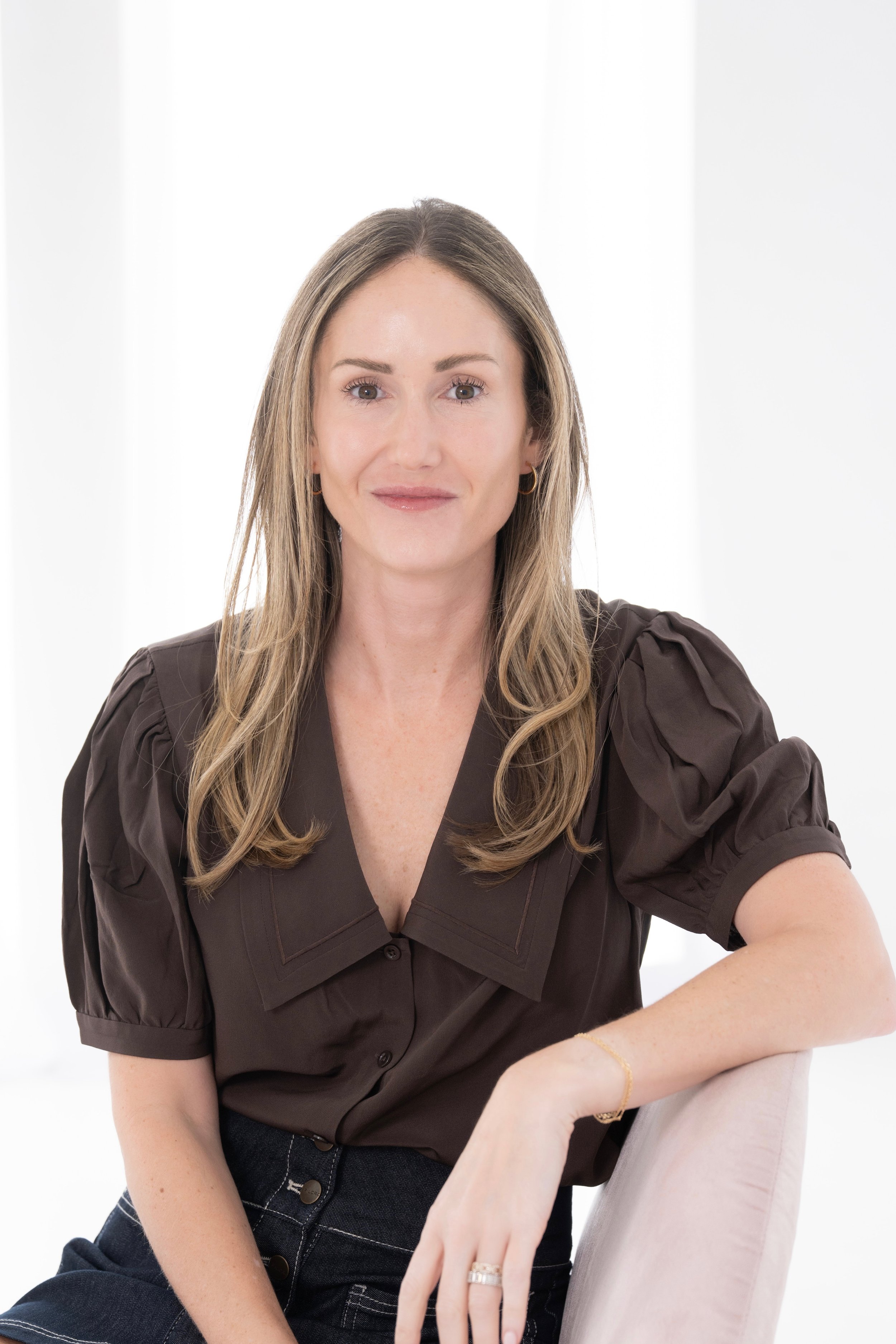 A woman with long blonde hair wearing a brown blouse with puffed sleeves sitting on a beige chair in a bright room.