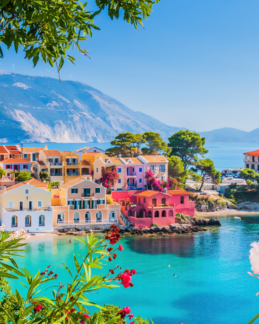 Colorful houses by a turquoise bay with mountains in the background, framed by green trees and pink flowers in the foreground.