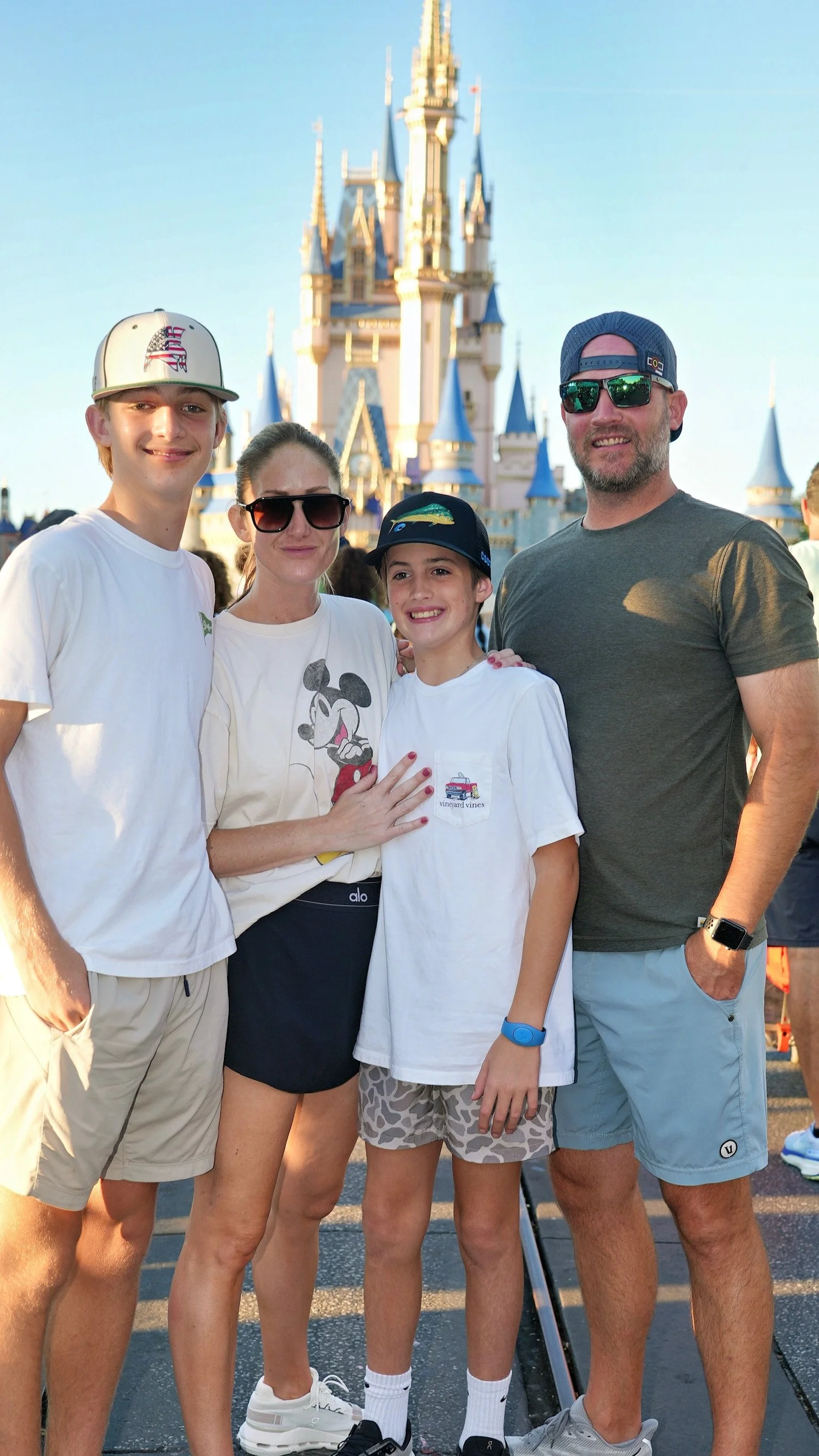 A family of four posing in front of Sleeping Beauty Castle at Disneyland, with a clear blue sky overhead.