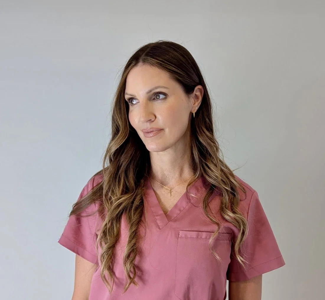 A woman with long wavy brown hair wearing pink medical scrubs and small hoop earrings looking to the side in front of a plain light-colored background.