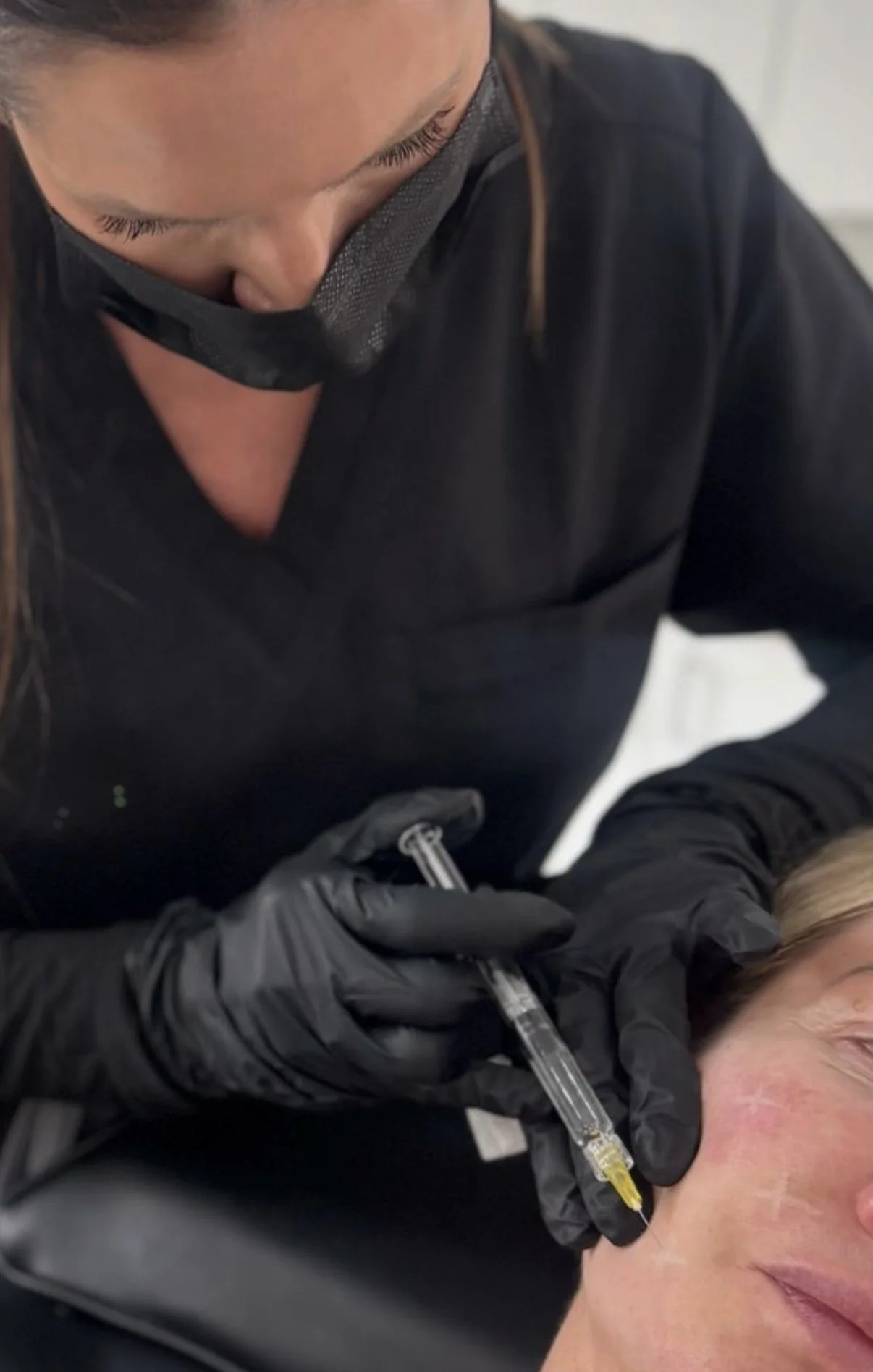 A woman receiving a cosmetic injection in her face from a professional wearing black gloves and a black mask.