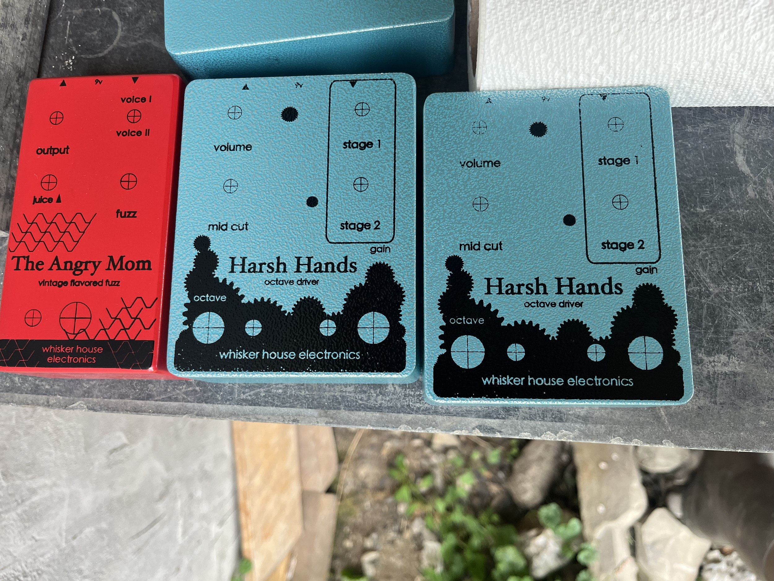 Two blue guitar effects pedals labeled Harsh Hands, with black text and knobs, placed on metal surface next to a red pedal labeled The Angry Mom with black text and graphics.