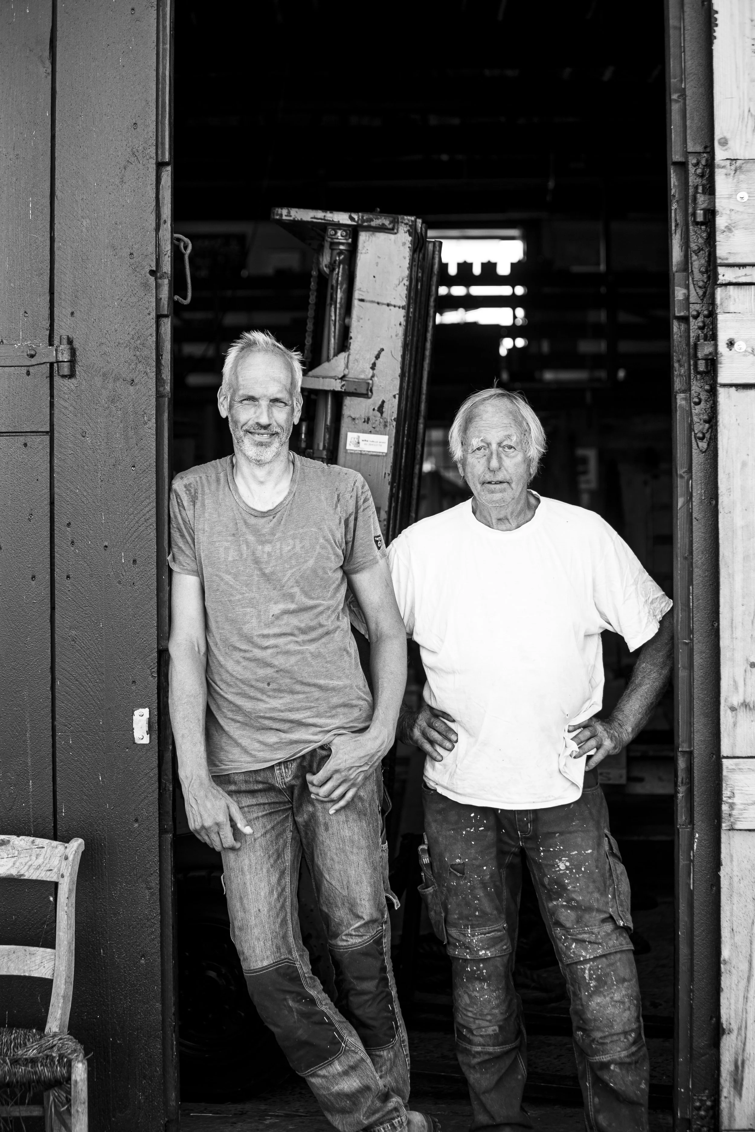 Twee mannen staan in de deuropening van een houten en metalen schuur. Ze lachen en kijken naar de camera.