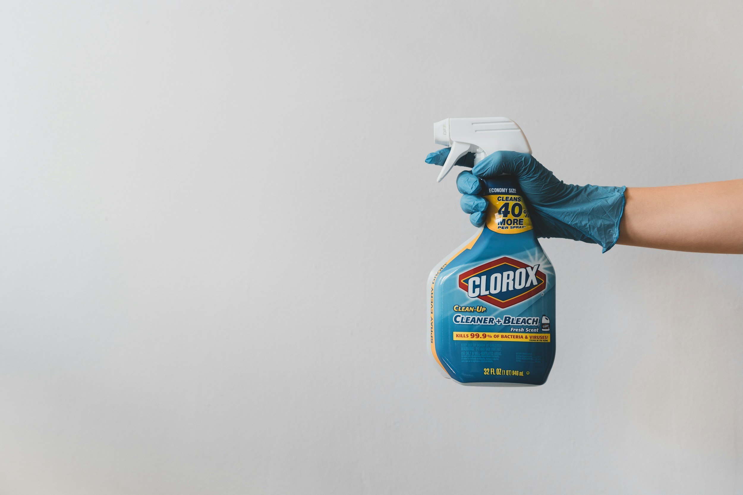 Hand wearing a blue glove holding a spray bottle of Clorox cleaner and bleach against a plain light-colored background.
