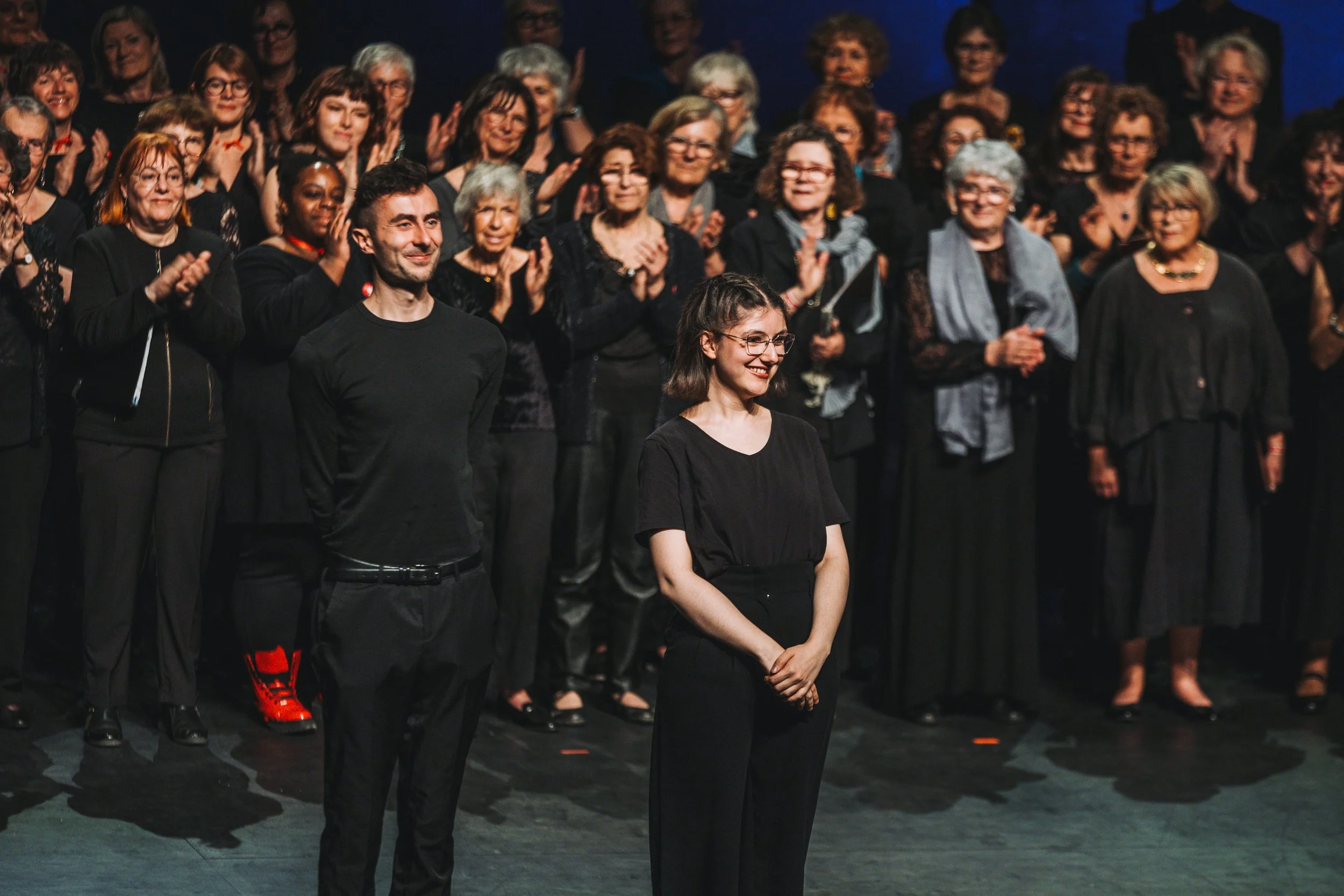 Un groupe de personnes sur scène, applaudissant et célébrant. Deux personnes se tiennent à l'avant, une femme et un homme, souriantes, avec une foule de femmes derrière eux, aussi souriantes et applaudissant.