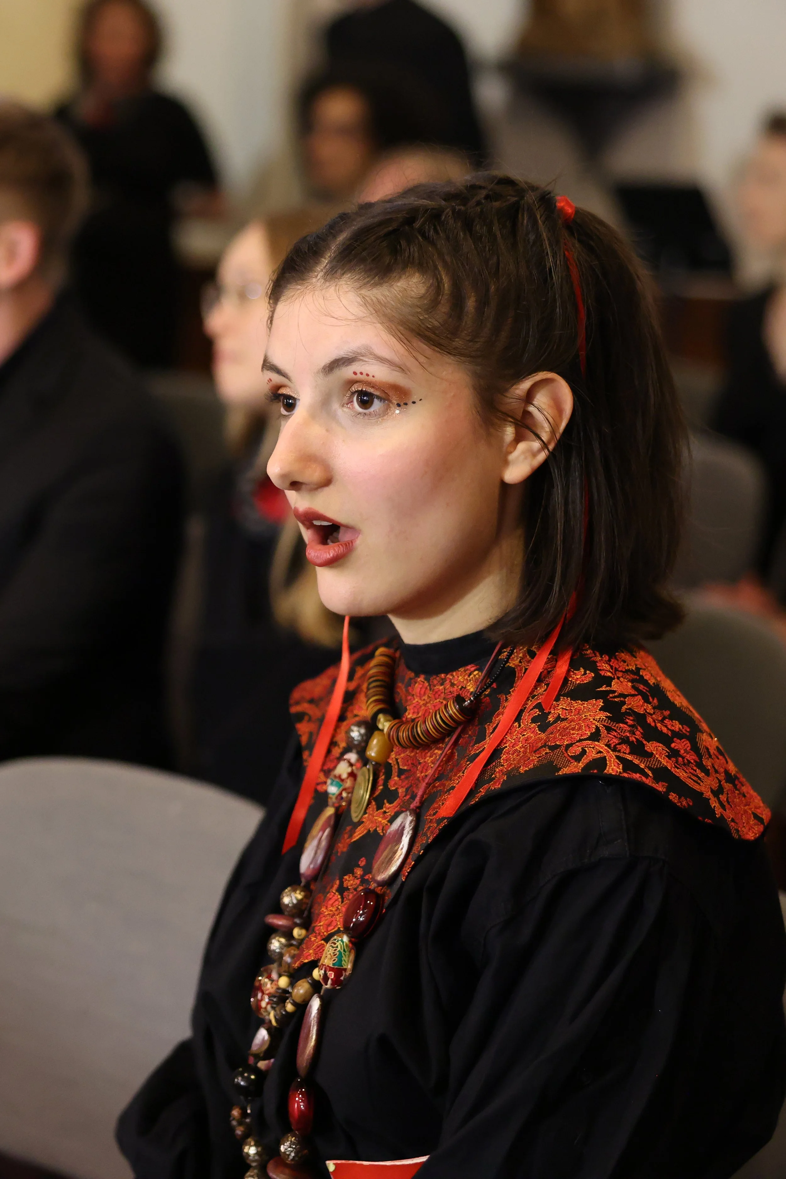 Une jeune femme avec des cheveux bruns mi-longs, portant un maquillage avec des accents de bijoux brillants autour des yeux. Elle porte une robe noire avec un col rouge orné d'un motif floral. Elle a également plusieurs colliers de bijoux colorés.