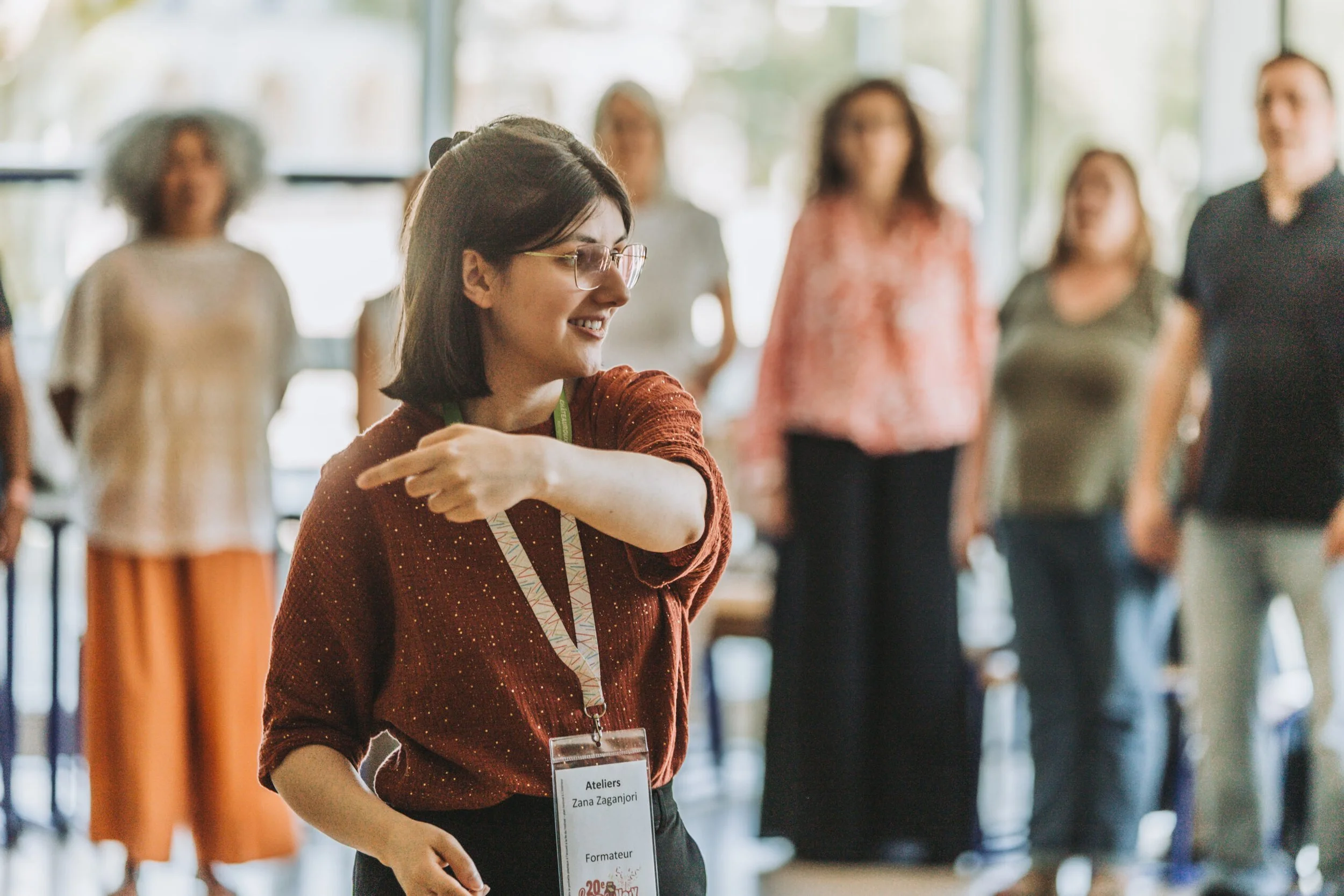 Une femme souriante portant un badge lors d'un atelier, avec d'autres personnes flou en arrière-plan dans une salle lumineuse.
