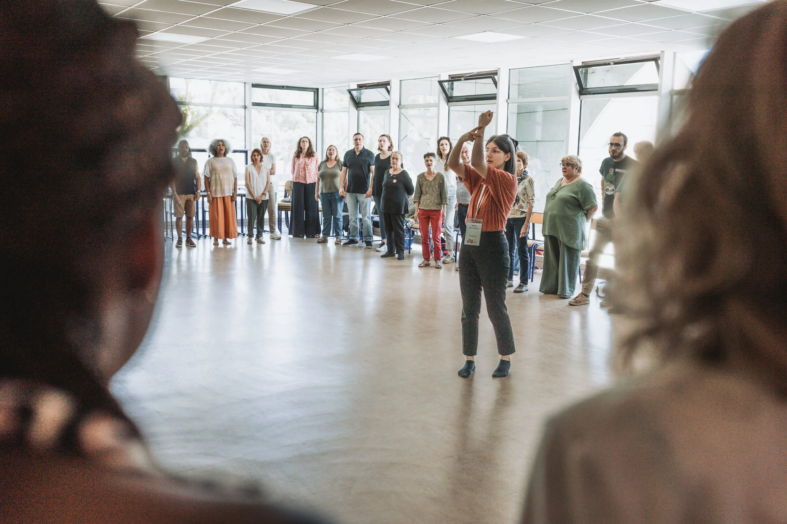 Un groupe de personnes assises et debout dans une pièce, observation une femme en train de faire une présentation ou une séance de chant, avec de grandes fenêtres en arrière-plan.