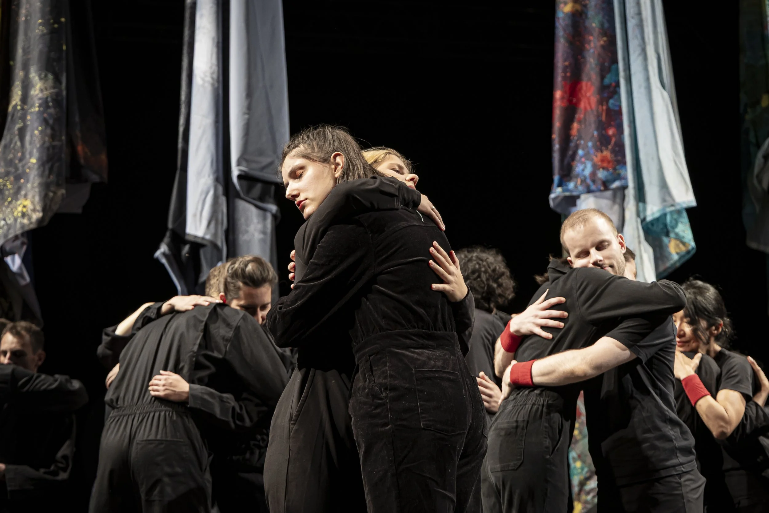 Groupe de personnes en noir s'embrassant sur scène, avec des vêtements suspendus en arrière-plan.