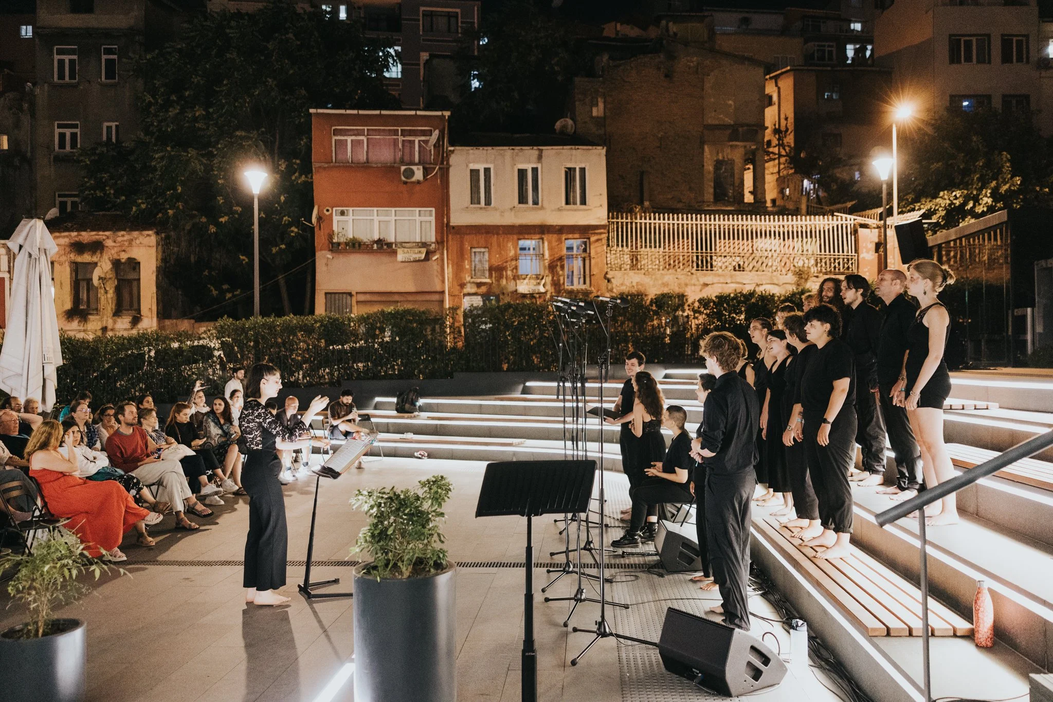 Une groupe de choristes en noir, dirigés par une chef d'orchestre, se produit sur une scène en plein air le soir, avec un public assis regardant la performance, dans un environnement urbain.