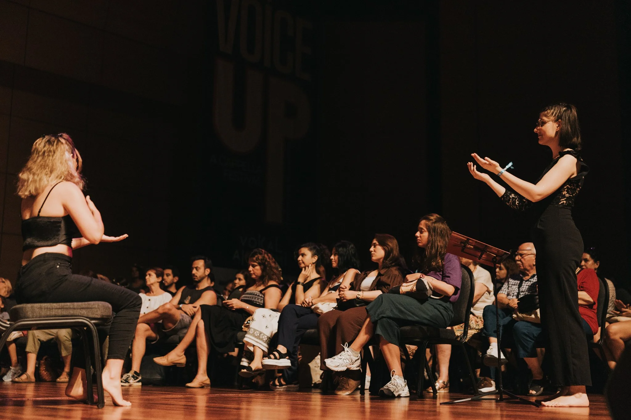 Une scène de théâtre ou de performance avec une femme en face de l'audience, qui est assise et regarde attentivement, dans une salle sombre. La femme semble parler ou performer, avec une autre femme debout, utilisant des gestes expressifs. L'audience est composée d'hommes et de femmes, certains prennent des notes ou regardent paisiblement.