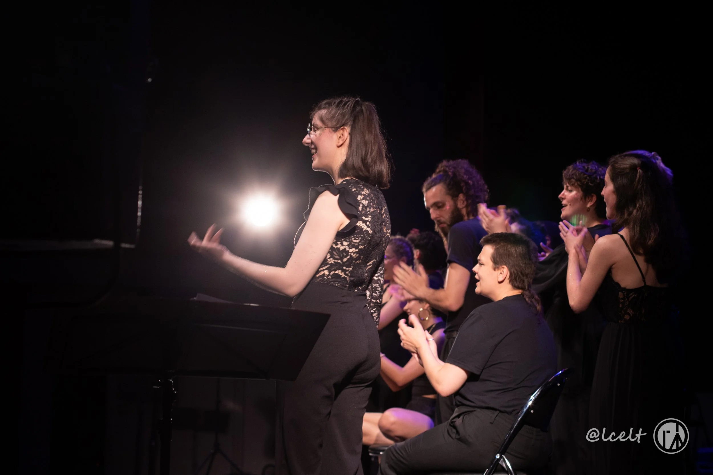 Groupe de personnes applaudissant et félicitant une femme sur scène lors d'une performance ou cérémonie, avec un éclairage de scène en arrière-plan. Il s'agit du choeur lyonnais VOX POP, dirigé par Zana Zaganjori.