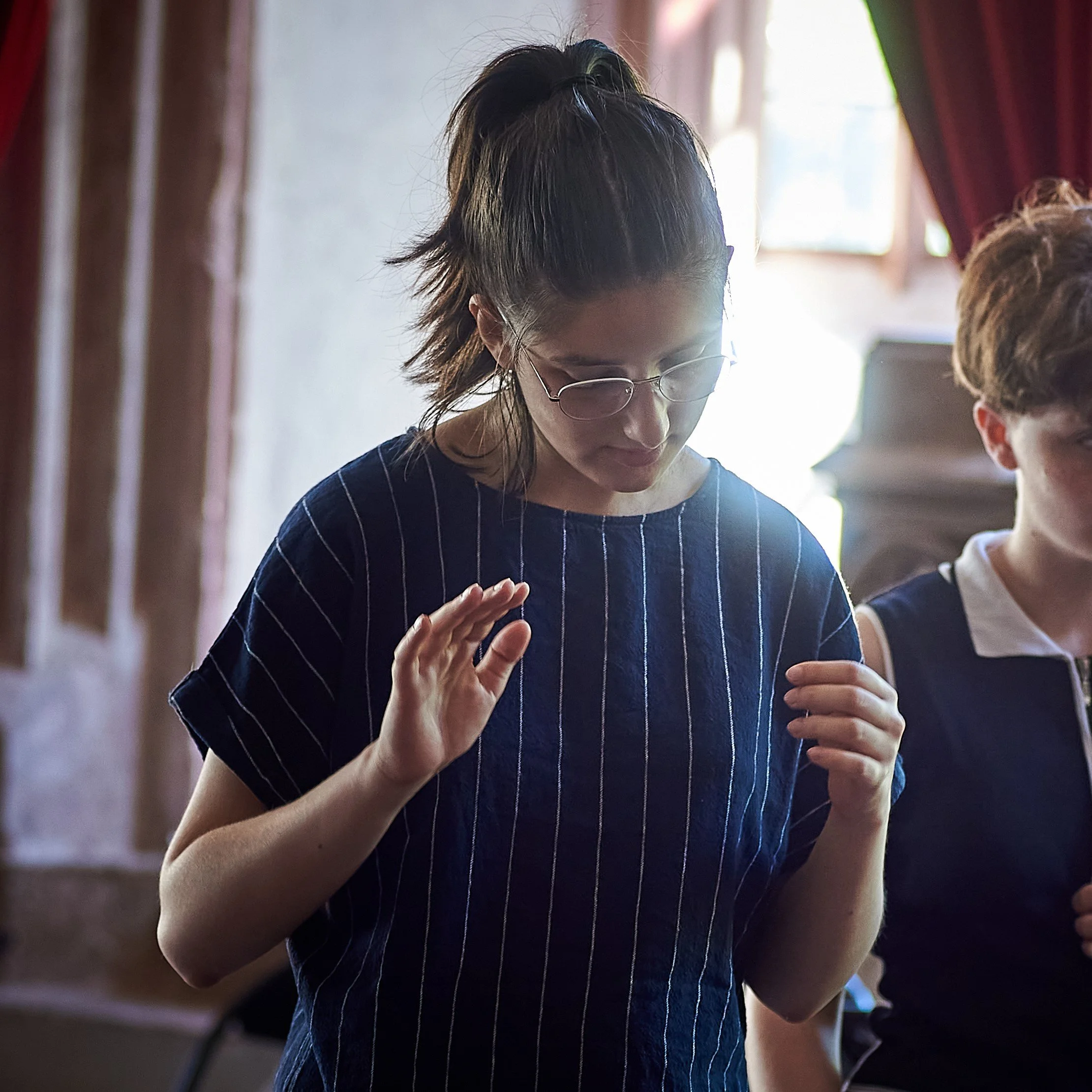 Une jeune femme avec des lunettes, portant un t-shirt à rayures, semble prier ou méditer dans une pièce éclairée par la lumière naturelle.