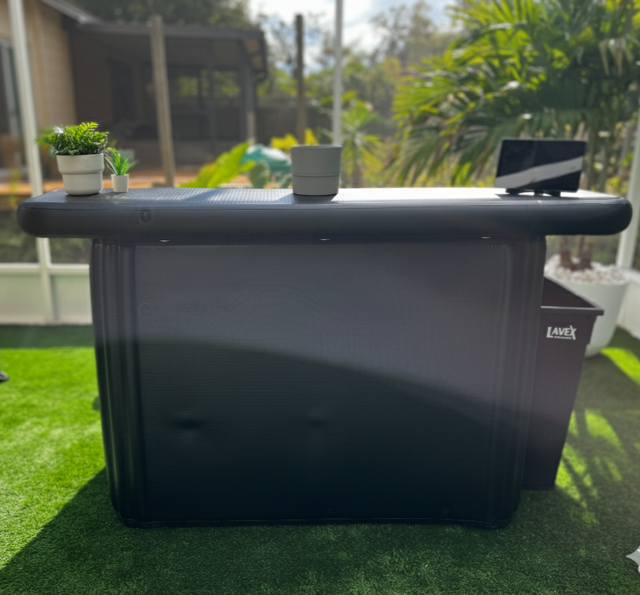 Outdoor space with a black storage container, potted plants, a plant pot, and a reflective tablet, with greenery and trees in the background.