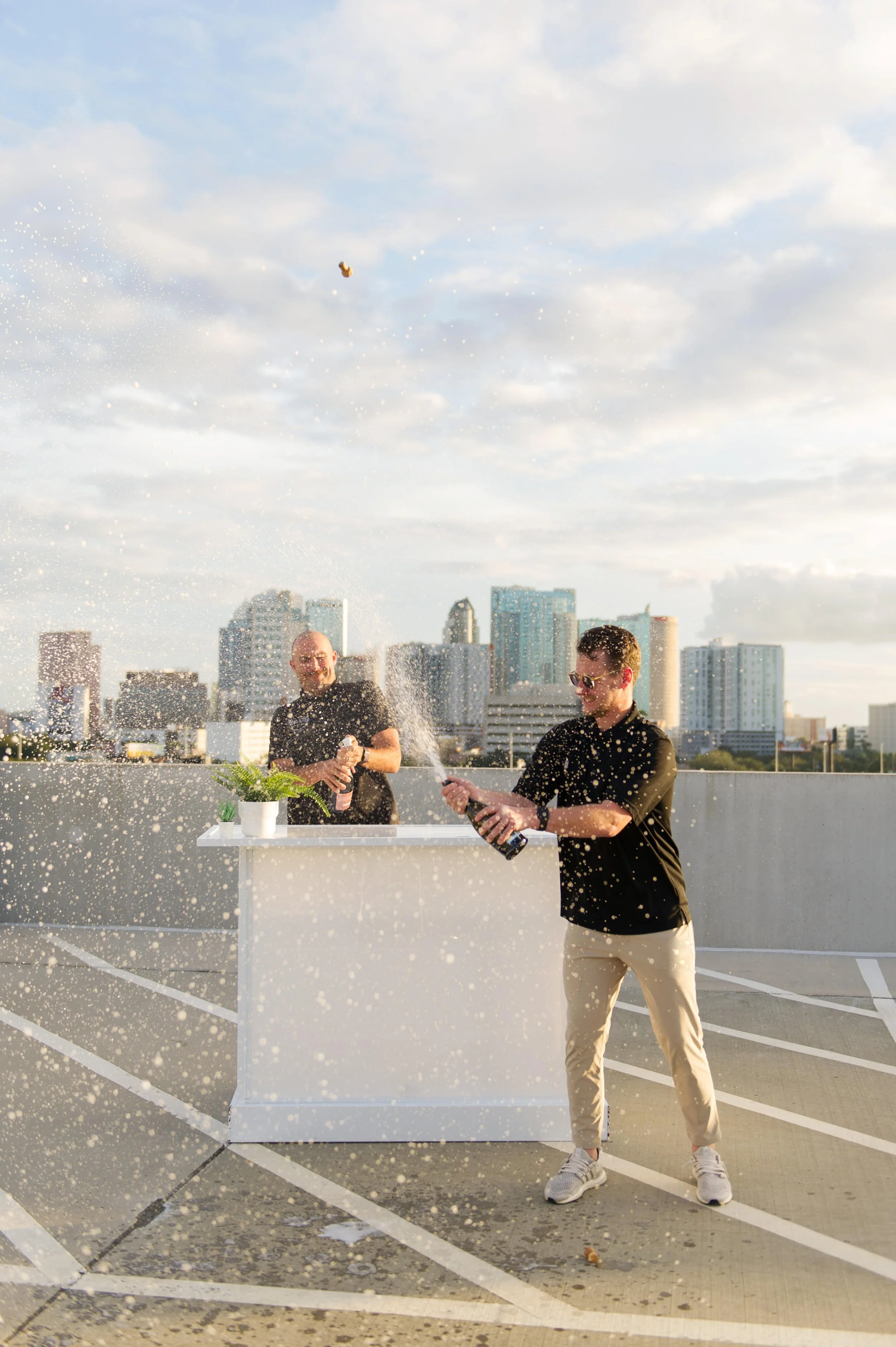 Two men celebrating with champagne on a rooftop parking lot with a city skyline in the background, one opening the champagne bottle while the other holds a bottle.