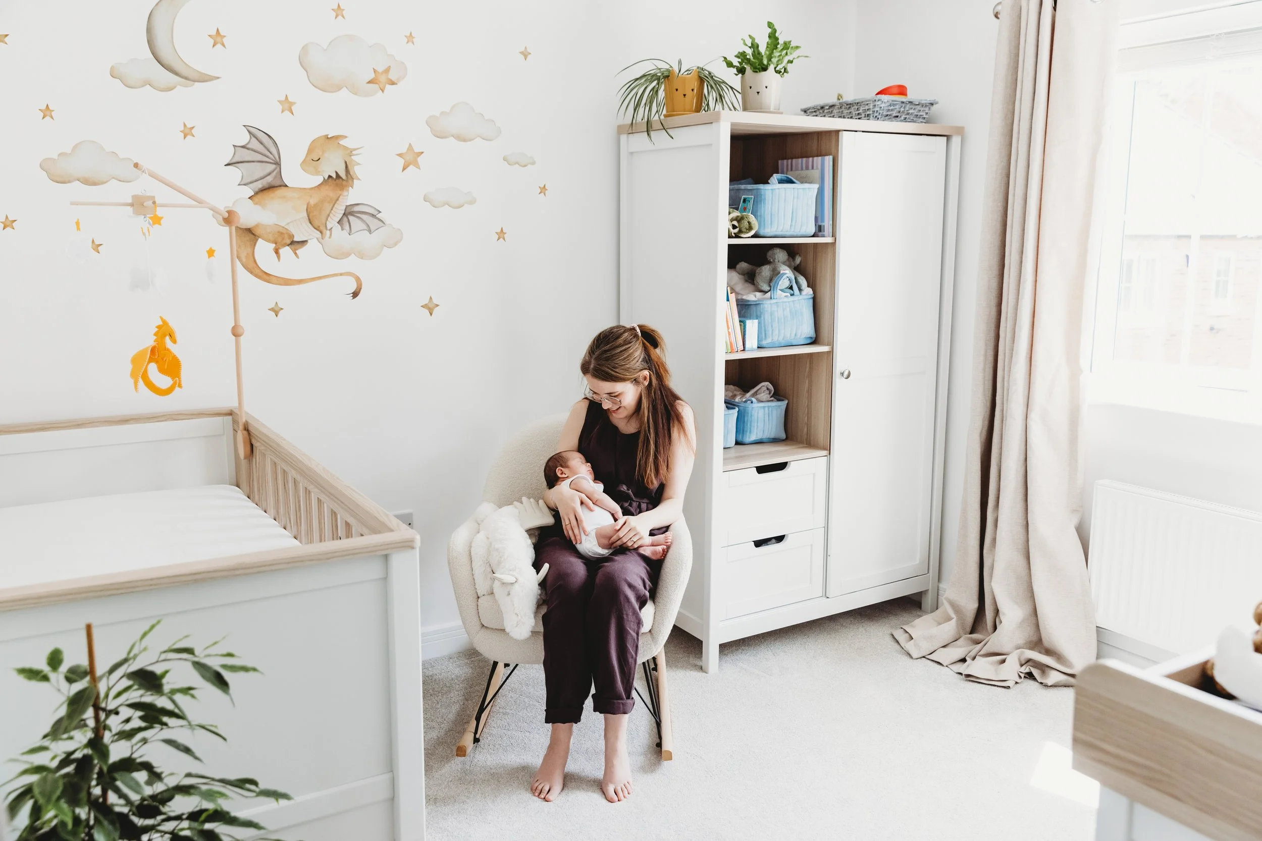 A woman sitting on a chair holding a newborn baby in a nursery with moon and cloud wall decorations, a wooden bed frame, a white closet with open shelves containing toys and books, a plant, and curtains near a window.