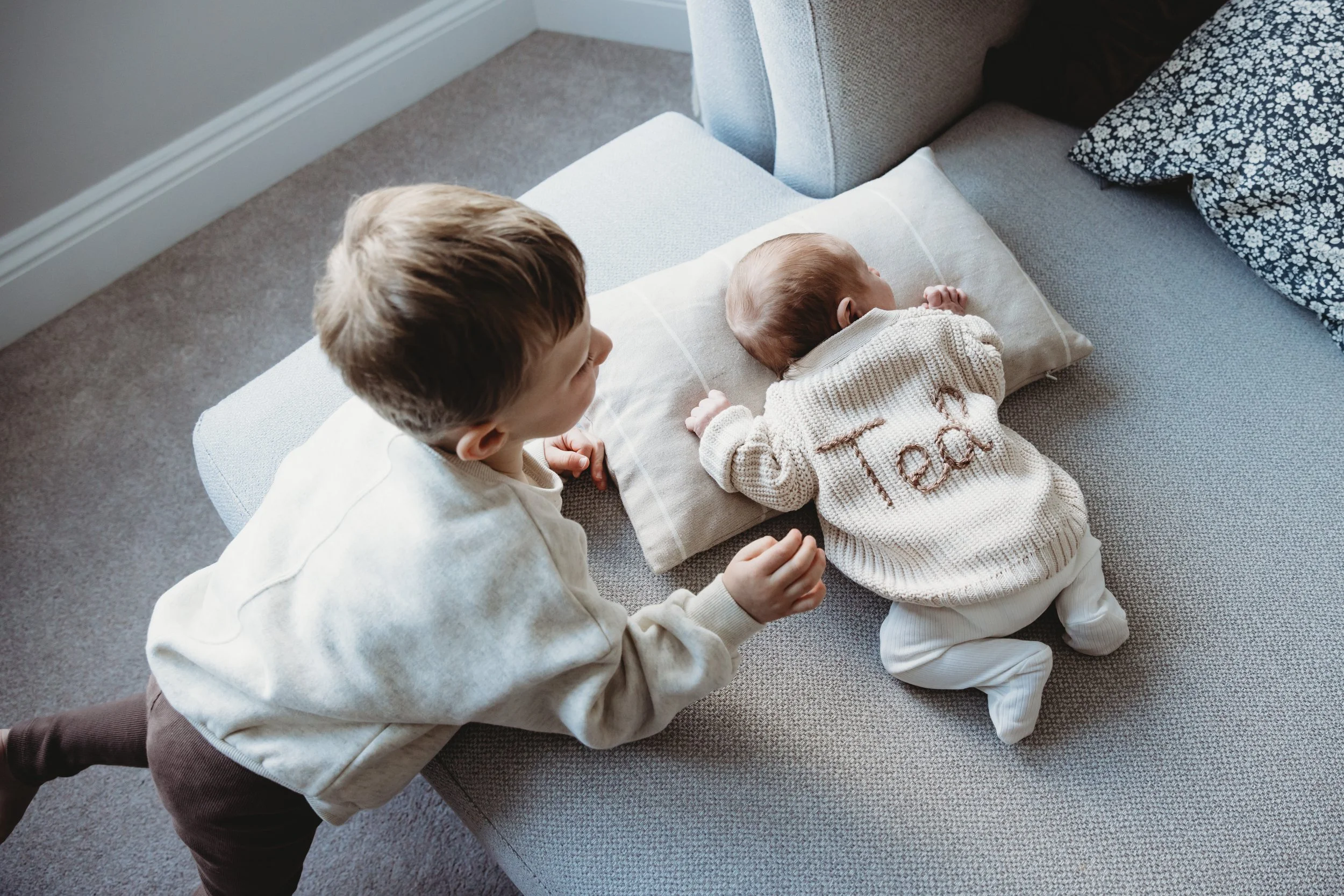 A young boy and baby lying on a couch, with the boy holding the baby's hand. The baby is on its stomach with its head turned to the side, and the boy is sitting nearby with a neutral expression.