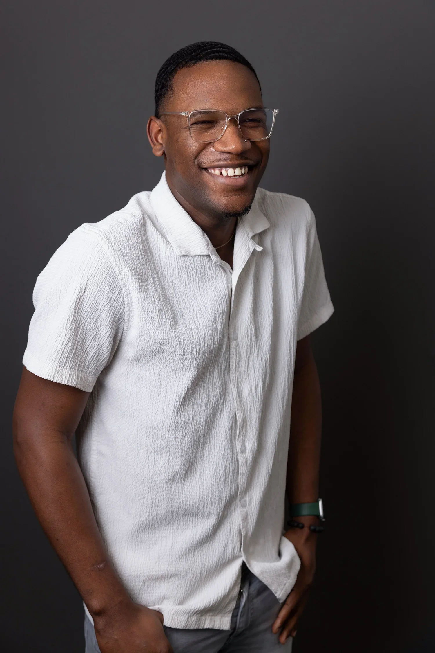 A smiling person with dark skin, wearing clear glasses and a white textured short-sleeve button-up shirt, standing against a dark grey background.