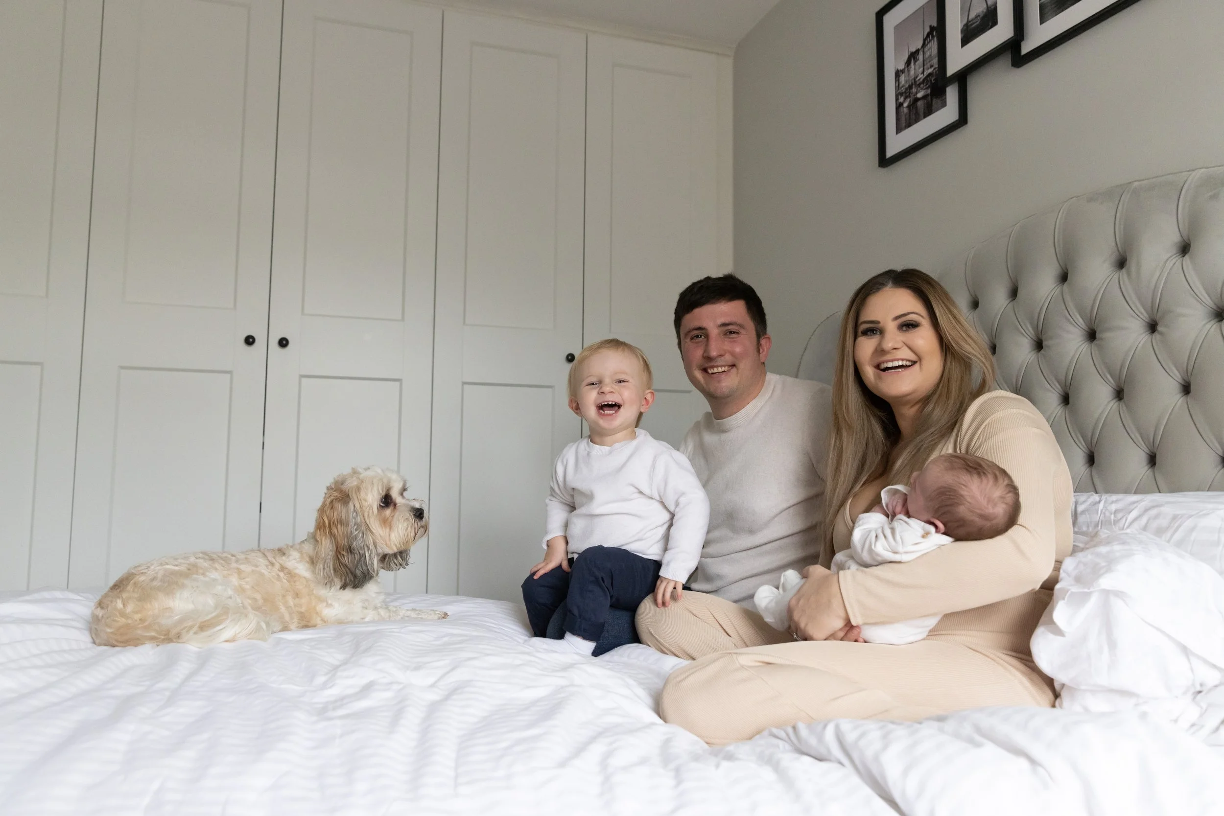 A family with two small children and a dog sitting on a bed in a bedroom with white walls and a tufted headboard, smiling and looking at the camera.
