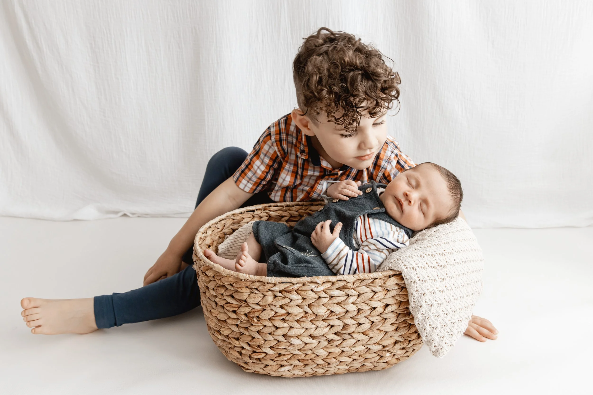 A young boy with curly hair leaning over and holding a sleeping baby in a basket, with a white backdrop.