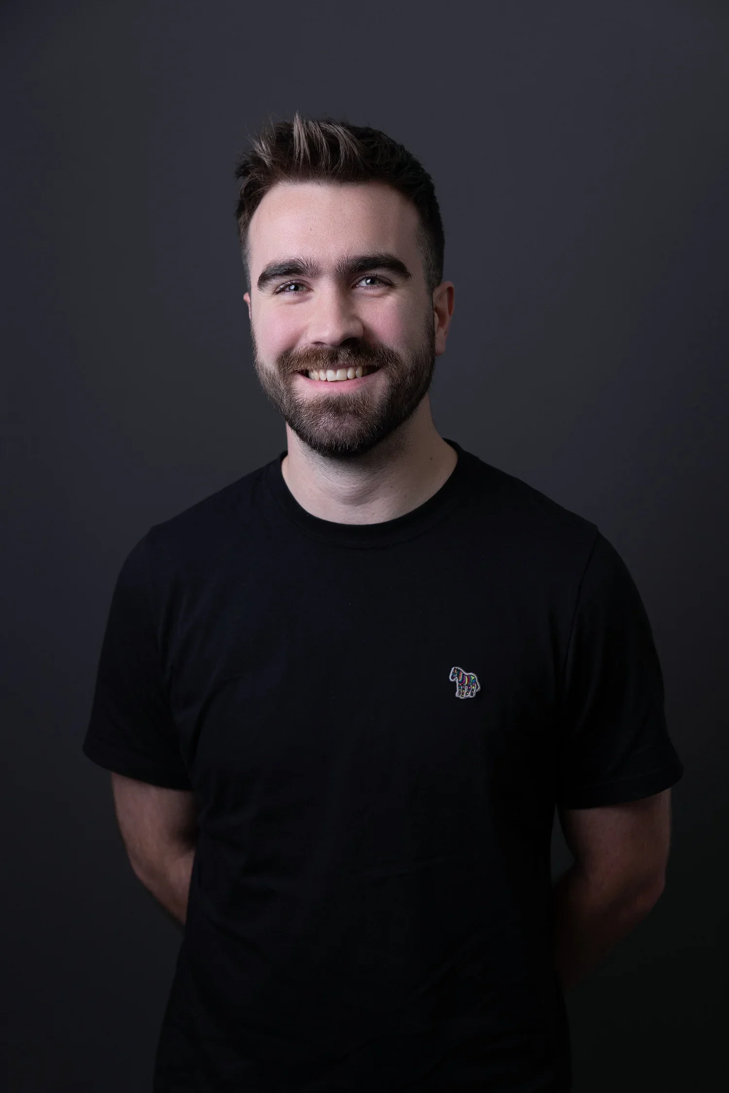 A young man with short brown hair and a beard, smiling, wearing a black t-shirt with a small colorful design on the chest, standing against a dark background.