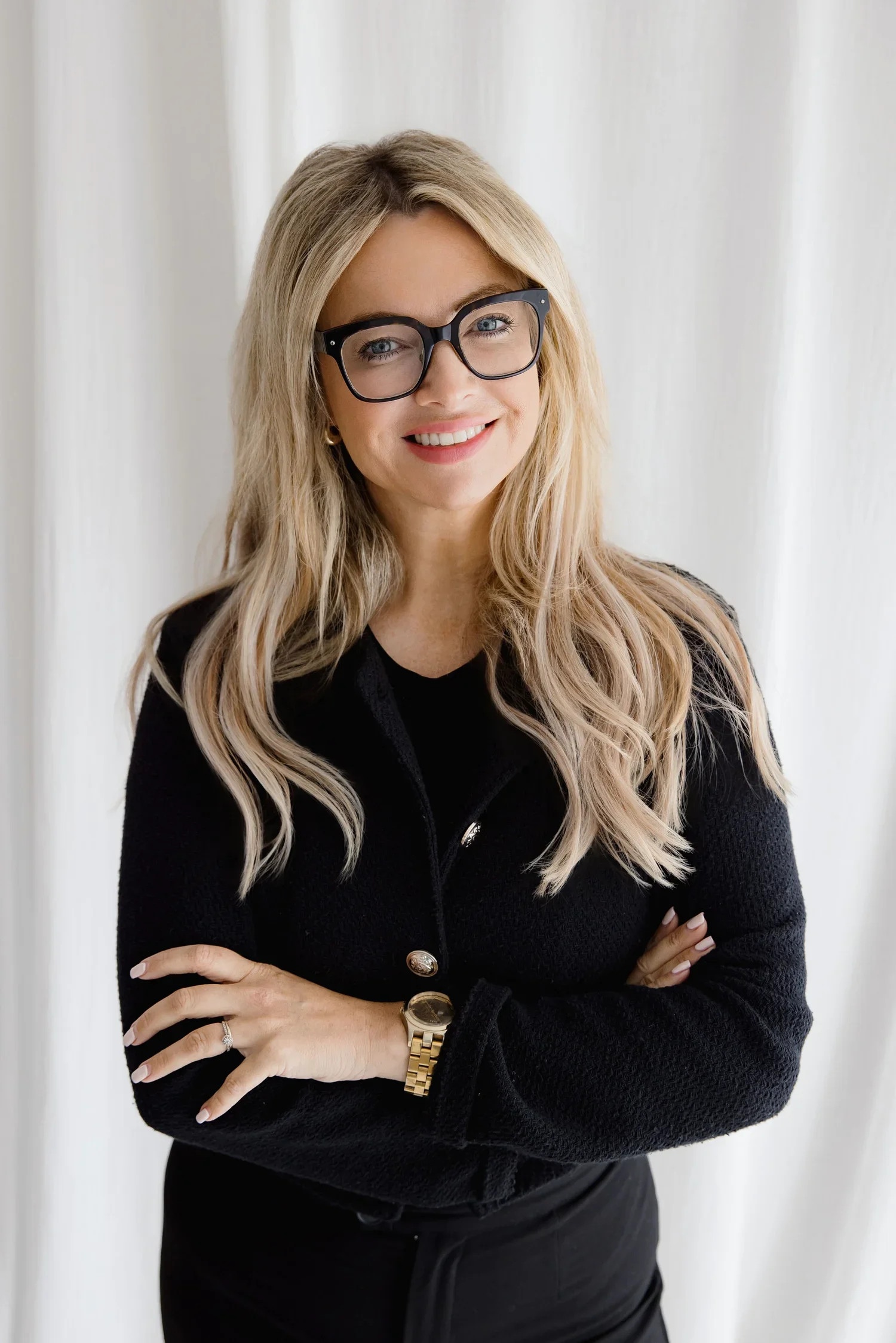 A woman with long blonde hair, wearing large black glasses, a black textured jacket with gold buttons, gold watch, and ring, smiling with arms crossed in front of a white curtain background.