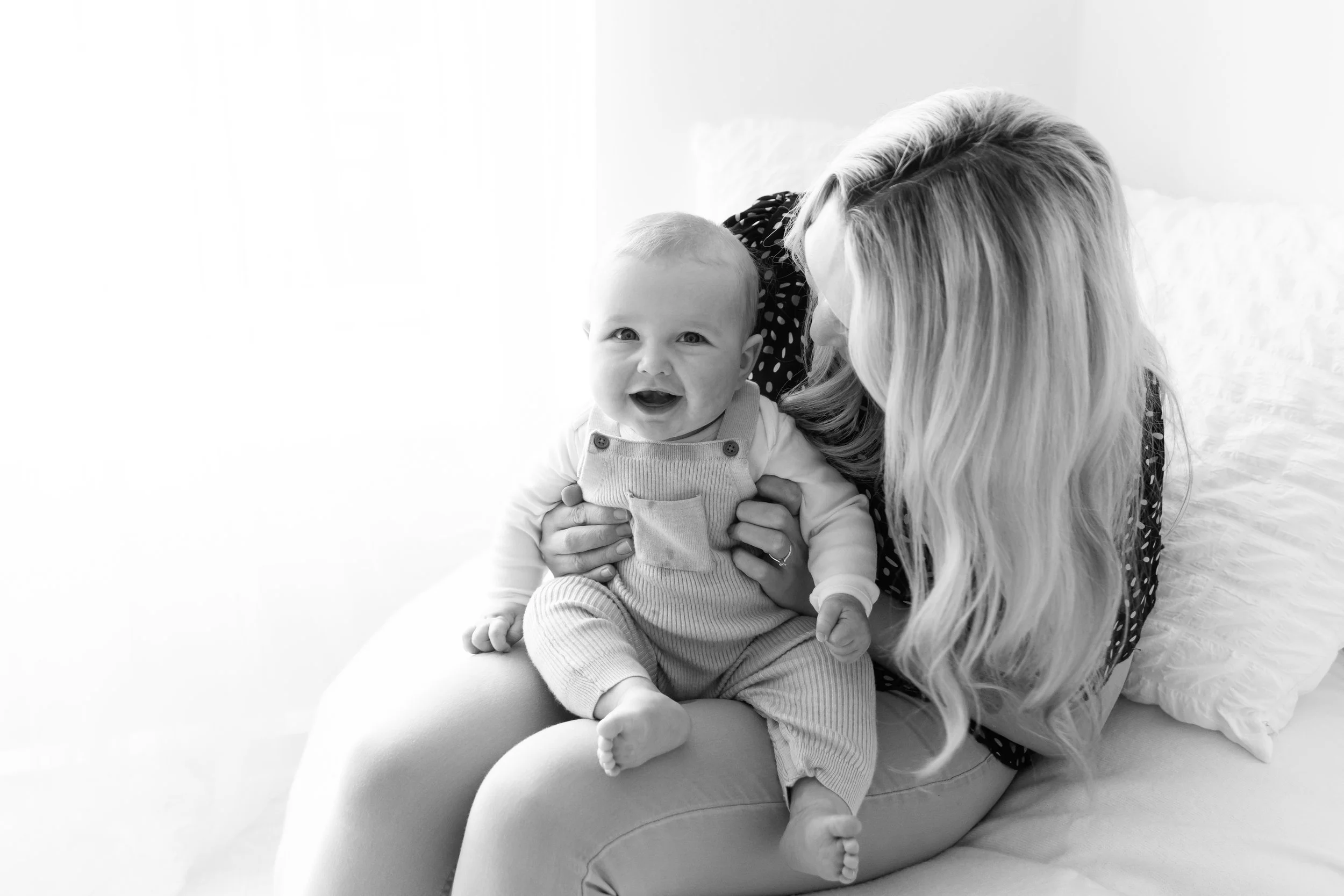 A woman with long blonde hair is holding and kissing a baby on the head. The baby is smiling and looking at the camera, wearing a light-colored outfit with overalls. They are sitting on a soft surface near a white wall.