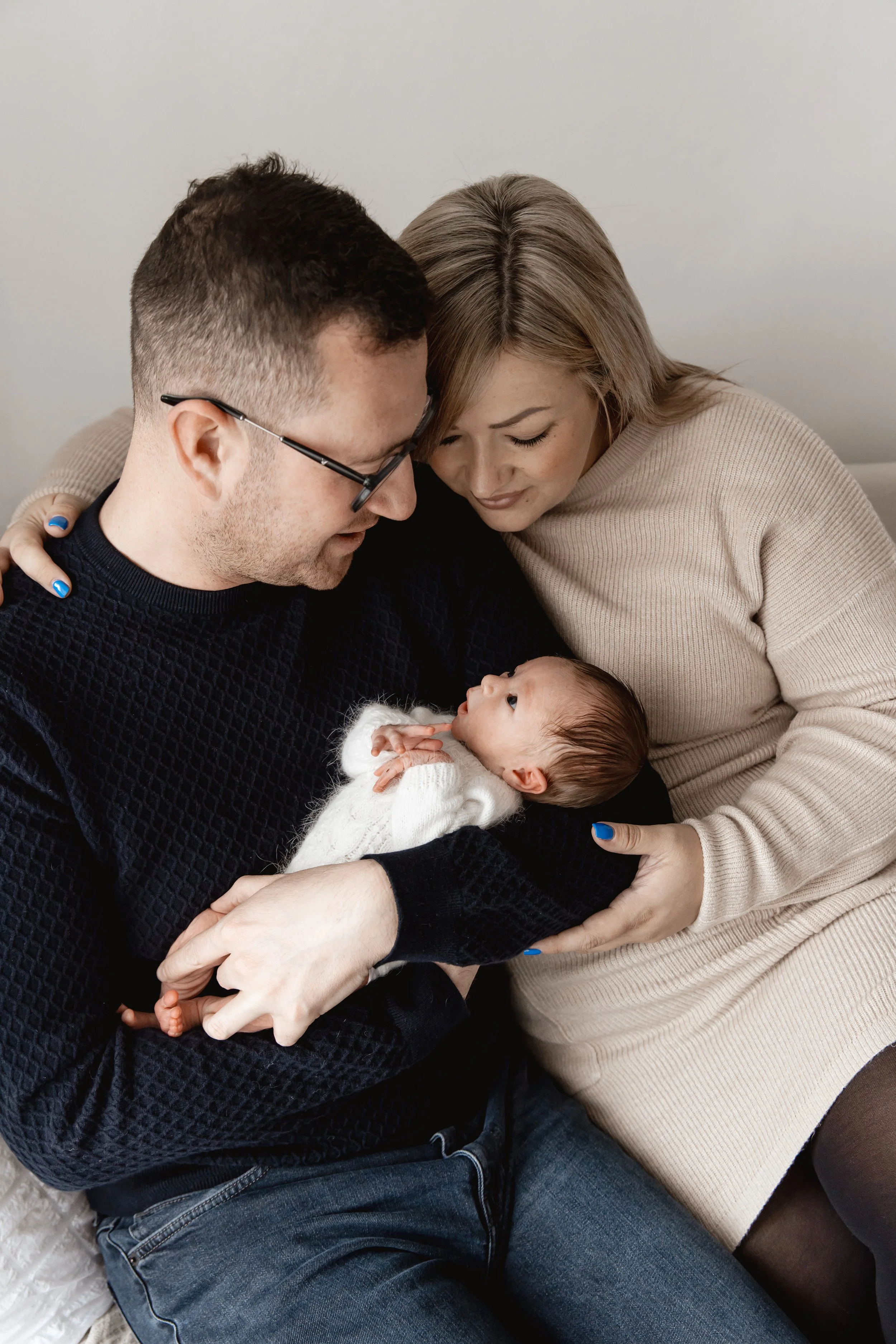 A family of three sitting on a couch, looking lovingly at their baby, who is lying on their laps. The father is wearing glasses and a dark sweater, the mother has blonde hair and is wearing a beige sweater, and the baby has dark hair and is dressed i