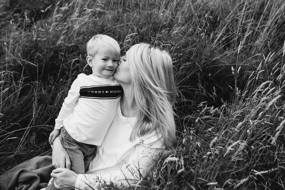 Black and white photo of a woman with long hair kissing a young boy on the cheek while sitting in a grassy field.
