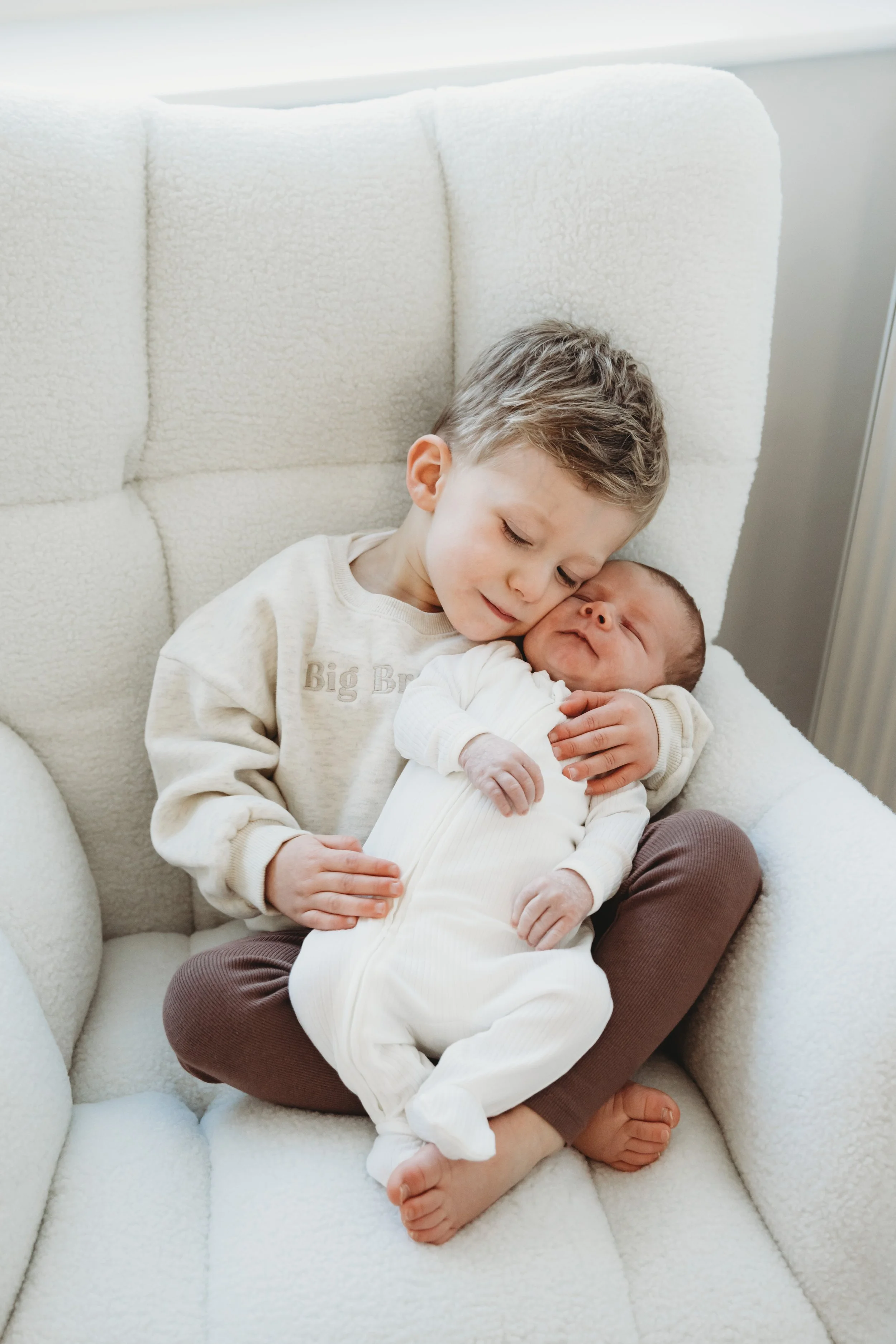 Two young brothers cuddling on a plush white armchair, with the older boy gently hugging the newborn while they sleep peacefully.
