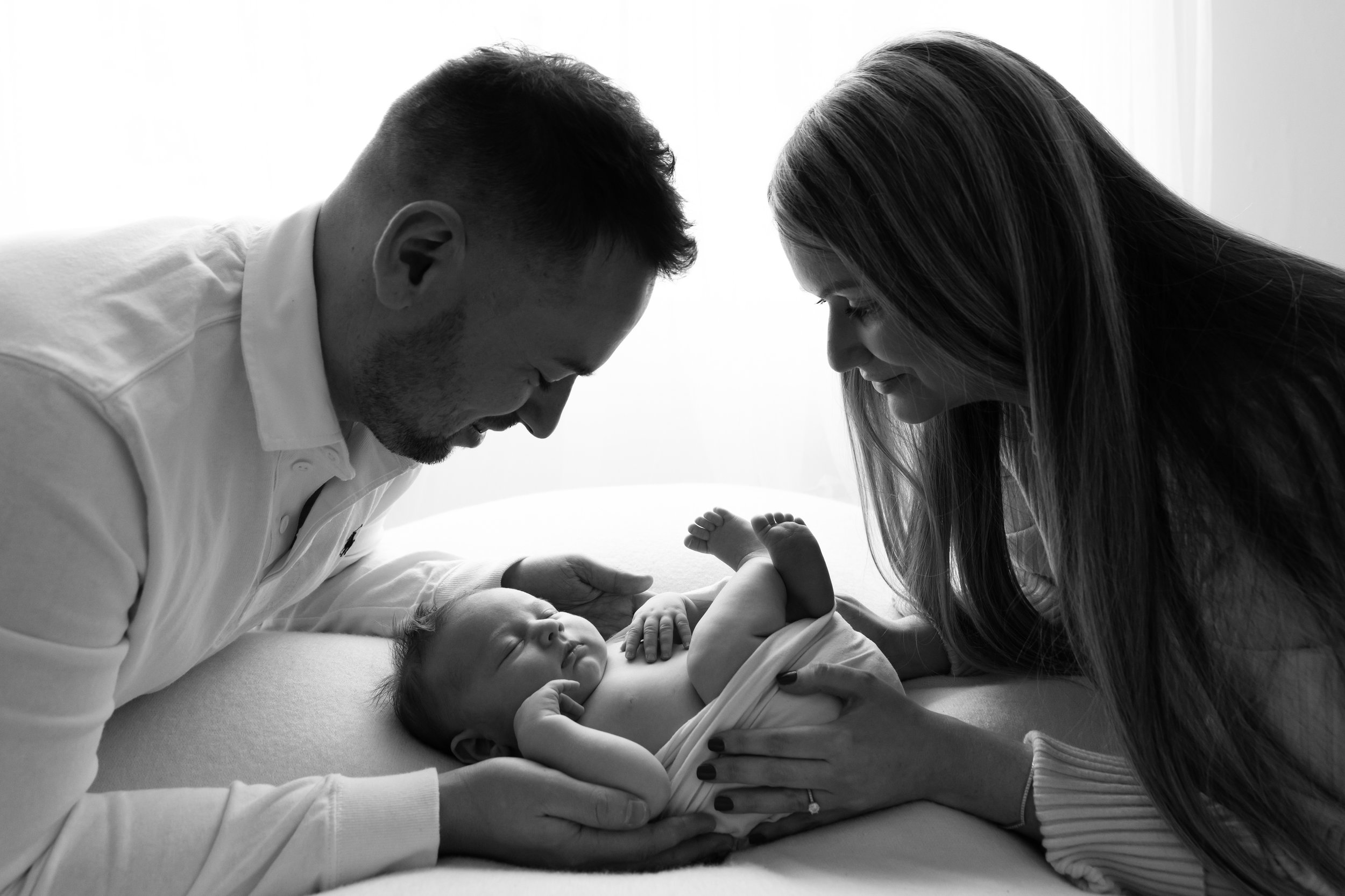 A black and white photo of a family in a hospital room. Two adults, a man and a woman, lean over a baby lying on a bed, with the woman gently touching the baby's arm and the man holding the baby's hand. The family appears to be happy and tender in th