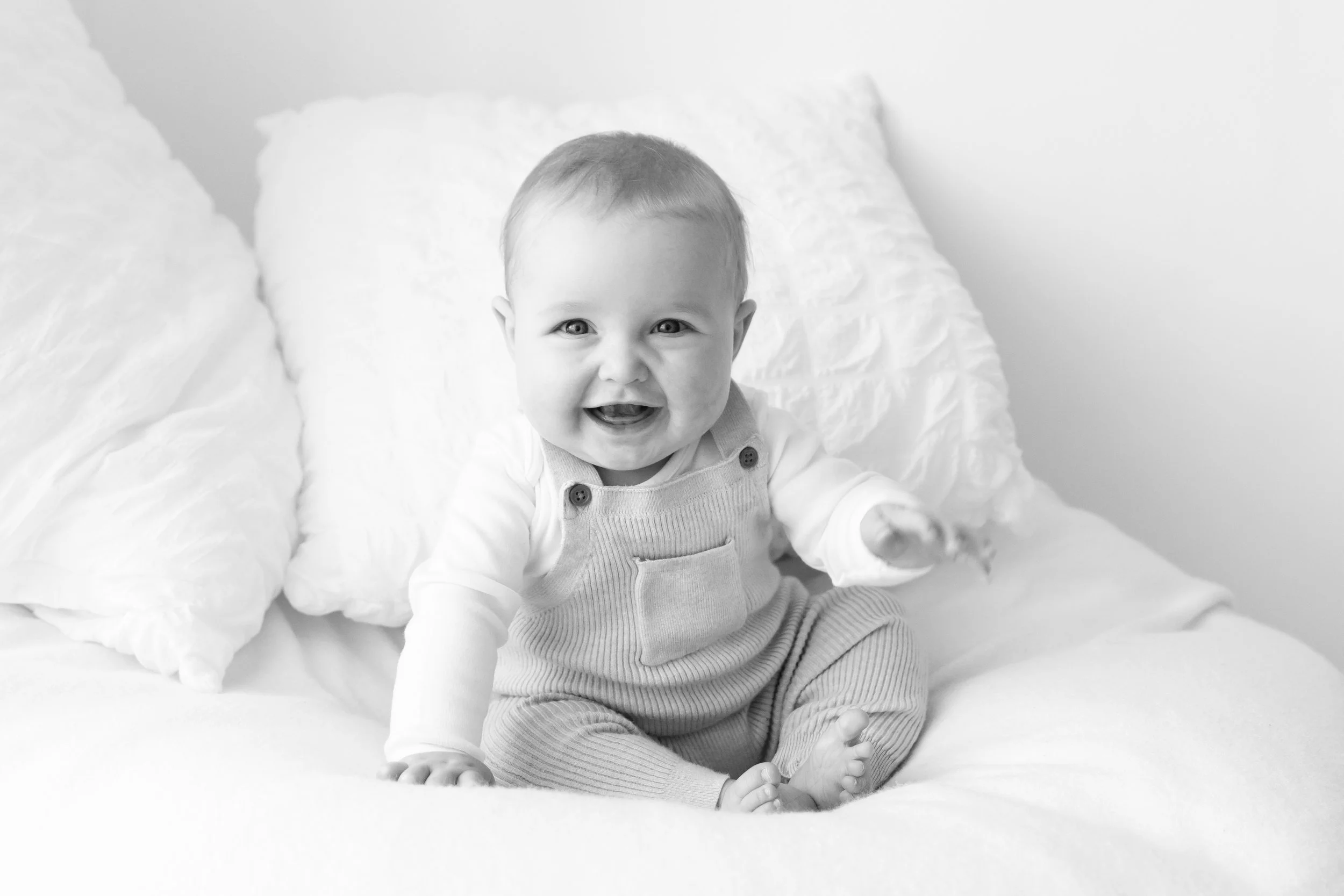 A joyful baby boy with light hair and bright eyes, smiling and sitting on a bed with fluffy pillows, wearing a striped outfit.