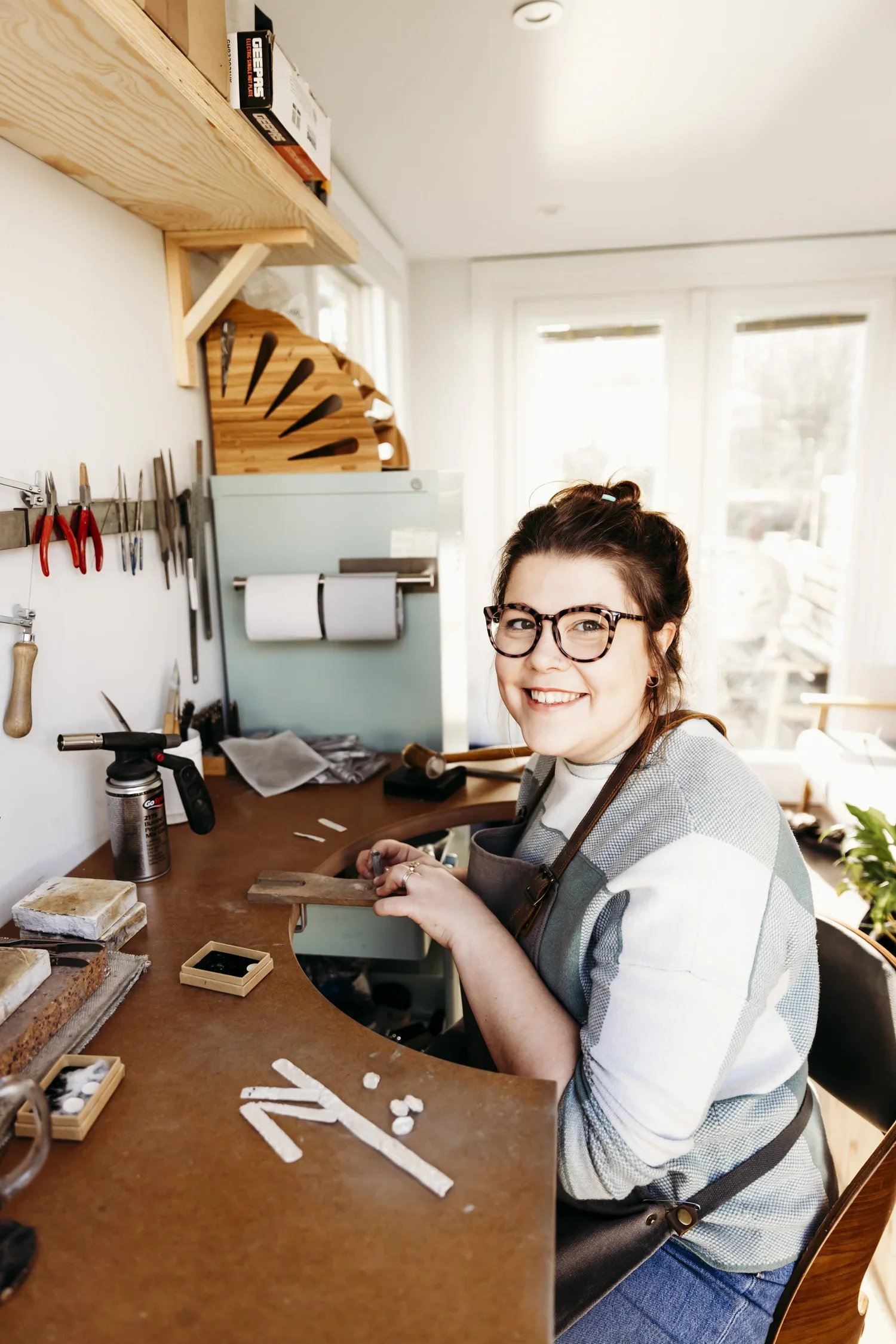 A woman with glasses and brown hair tied up, smiling at the camera, working at a wooden craft table with various crafting materials, in a bright room with large windows.