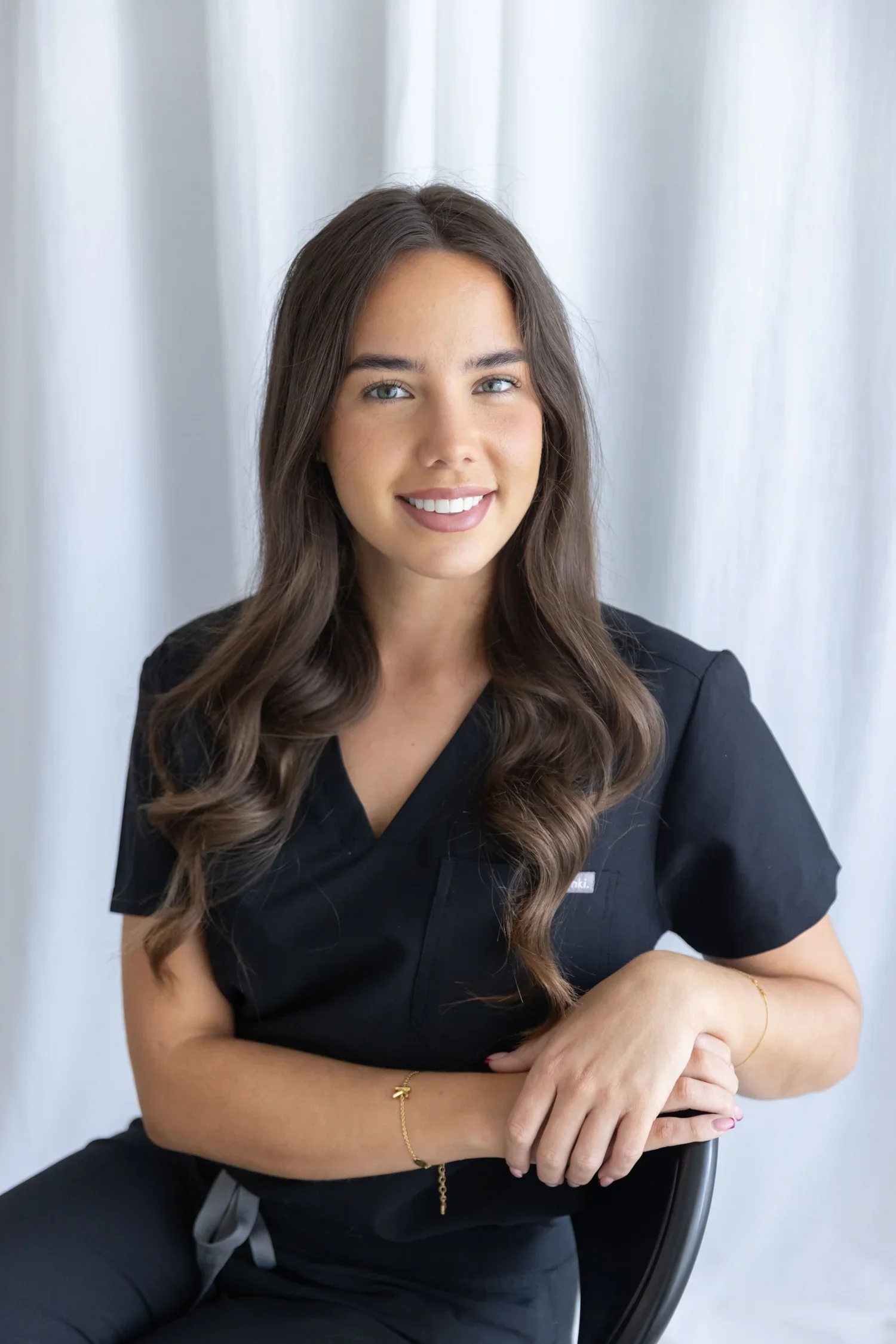 Headshot of a young woman with long wavy brown hair, blue eyes, and light skin, smiling while seated with crossed arms in front of a white curtain background.