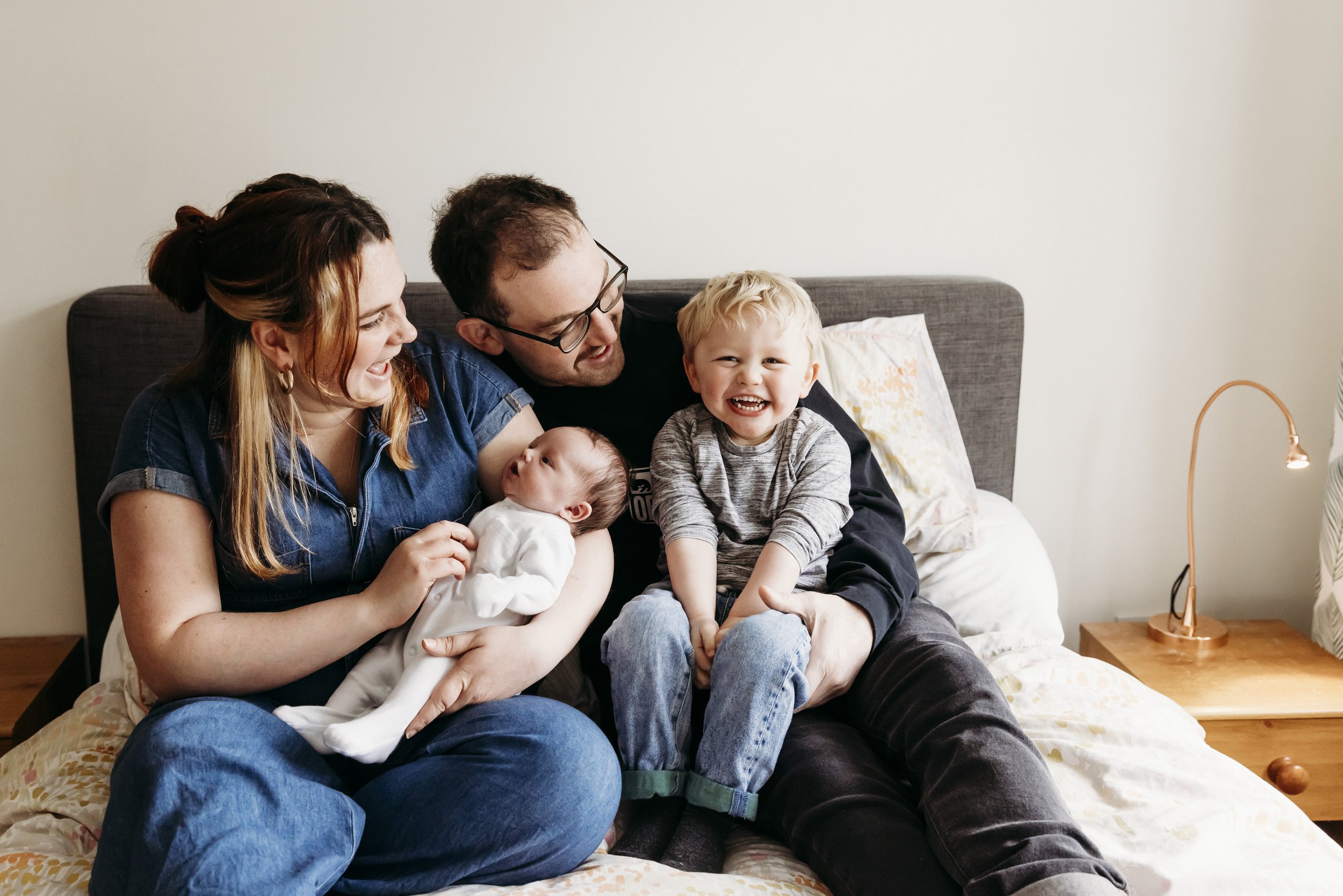 A family of five sitting on a bed and smiling, with a woman holding a baby, a man, a young boy, and a small child, in a bedroom with a gray headboard, a lamp, and pillows.