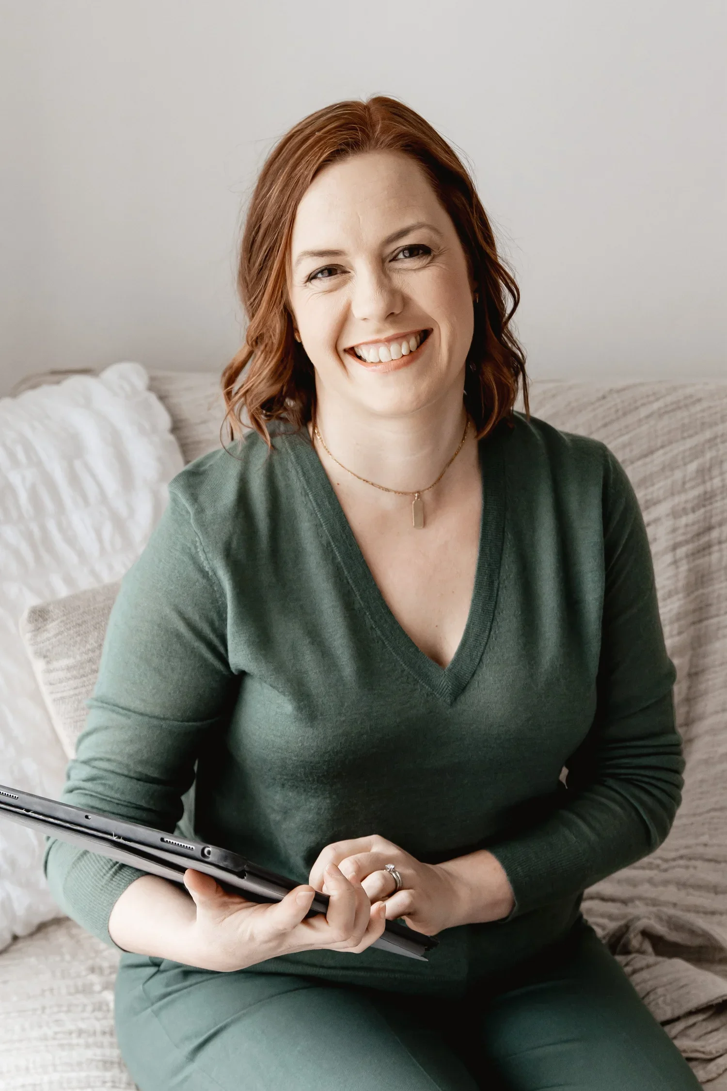A woman with red hair wearing a green sweater and holding a tablet, sitting on a beige couch.