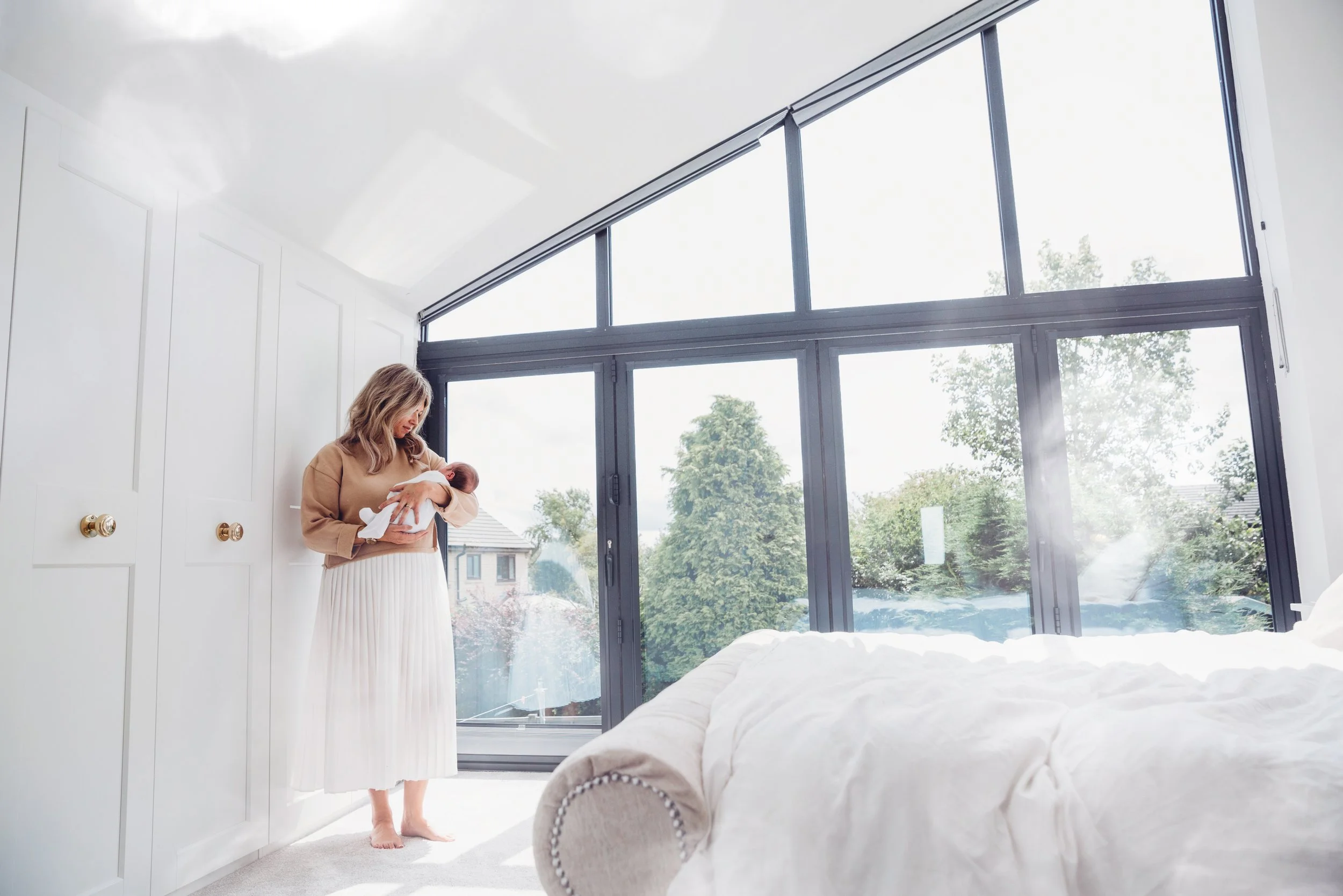 A woman holding a newborn baby inside a sunlit room with large windows showing trees outside, and white furniture.