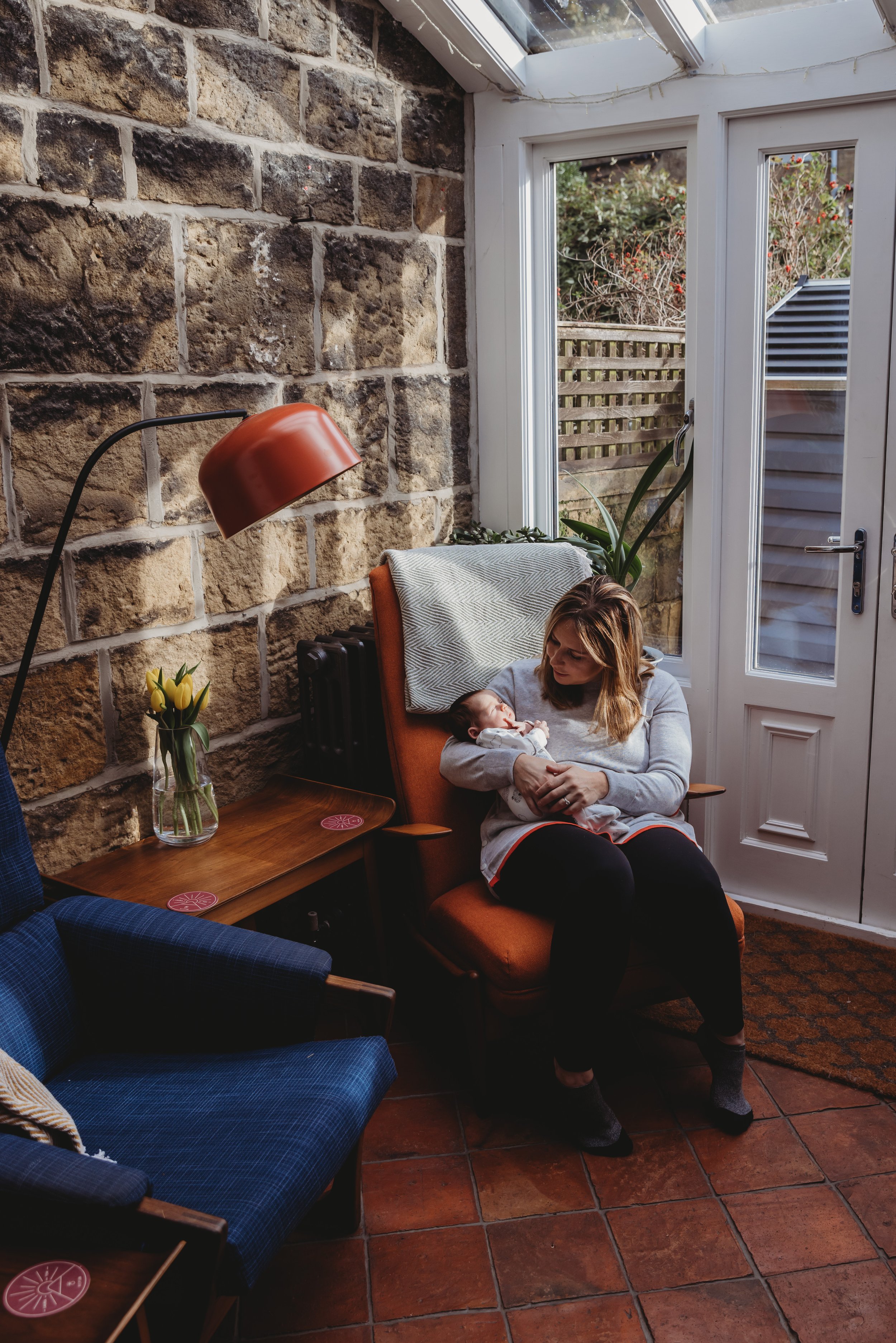 A woman sitting on a reddish-orange armchair in a sunlit room, breastfeeding a baby. The room has a stone wall, a glass ceiling, and large windows with white frames. There is a small table with a vase of yellow tulips nearby, and a blue checkered cha