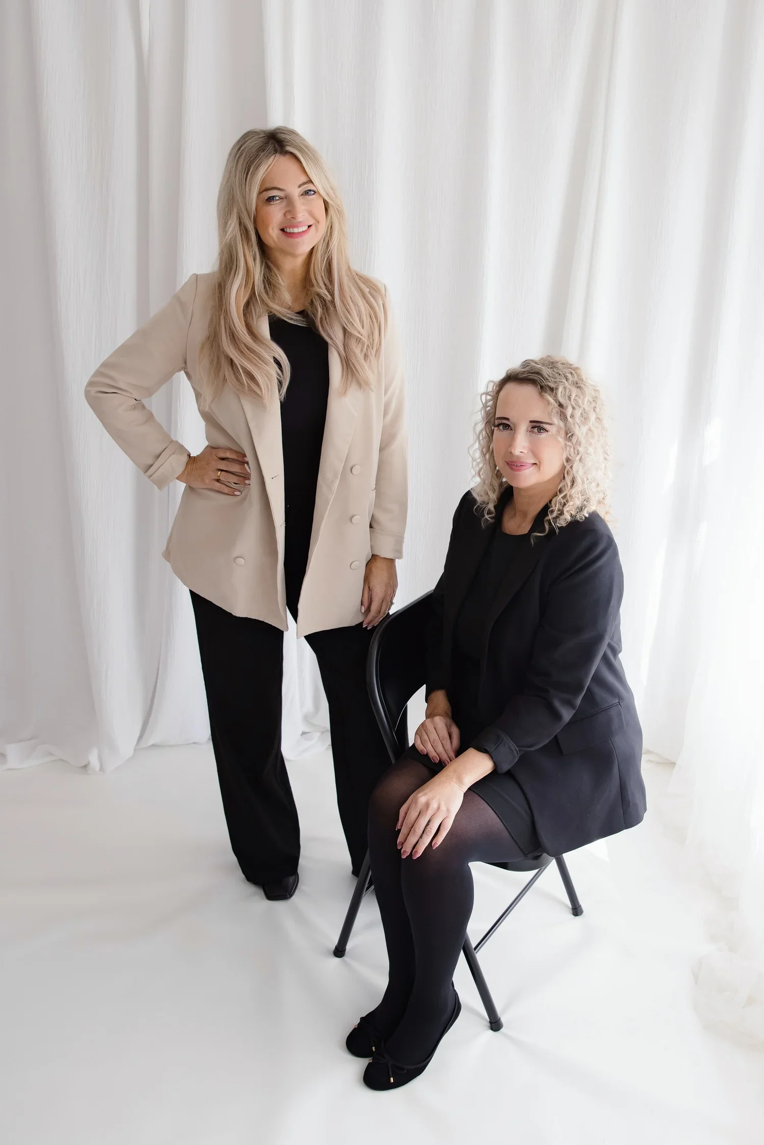 Two women posing for a photo in front of a white curtain, one standing and one seated on a chair, both dressed in black and beige professional attire.