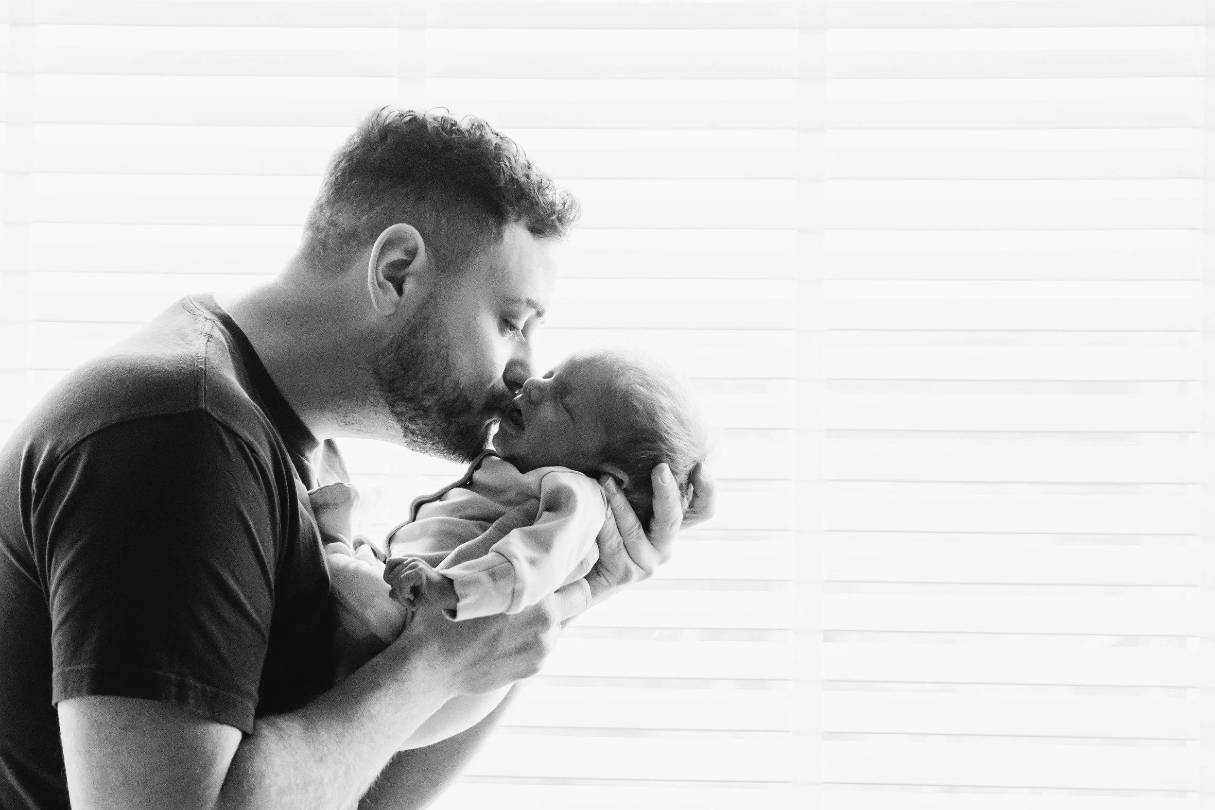 A man with a beard and curly hair holding a baby close to his face, both with closed eyes, in front of window blinds.