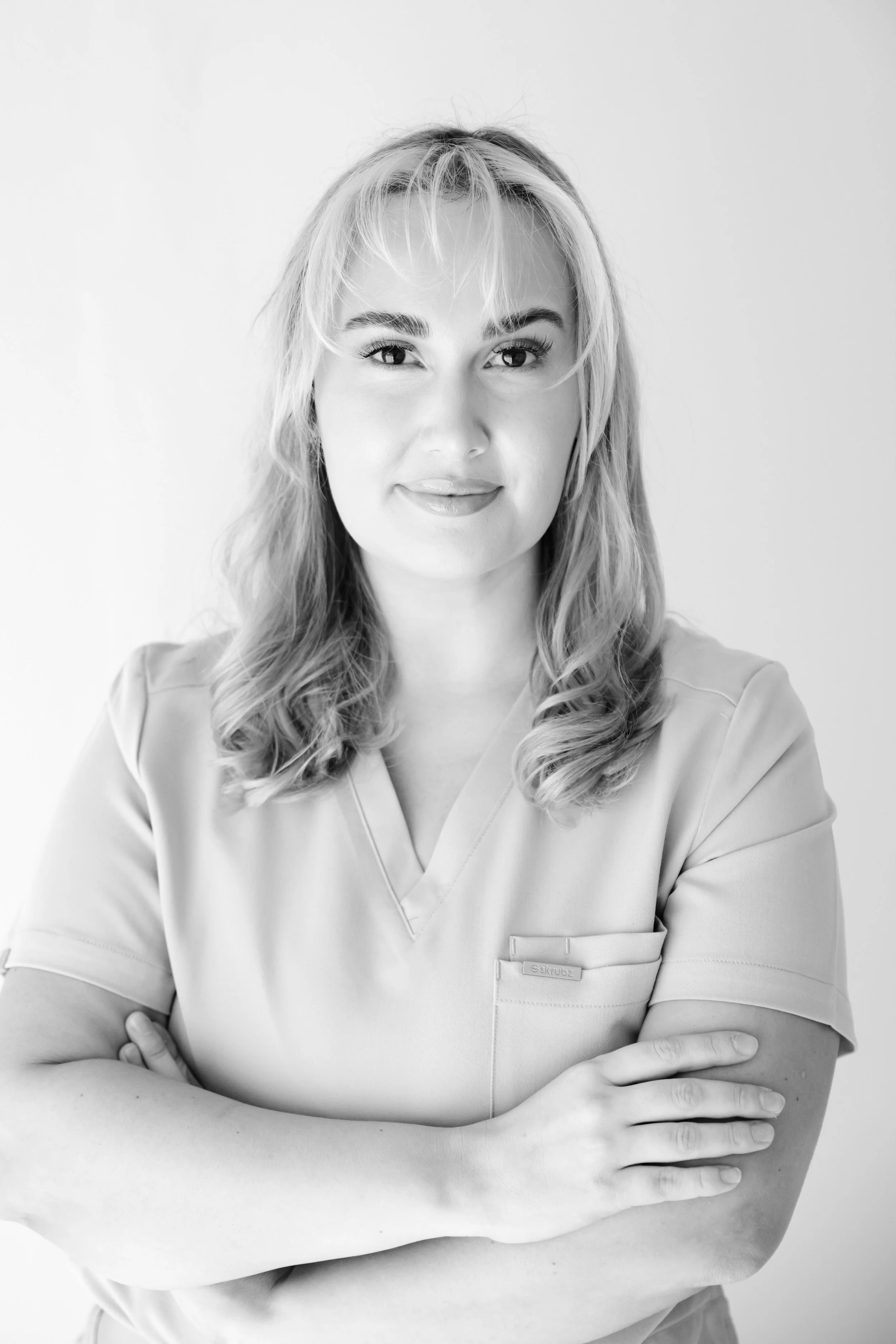 Black and white photo of a woman with blonde hair wearing medical scrubs, smiling softly with arms crossed.
