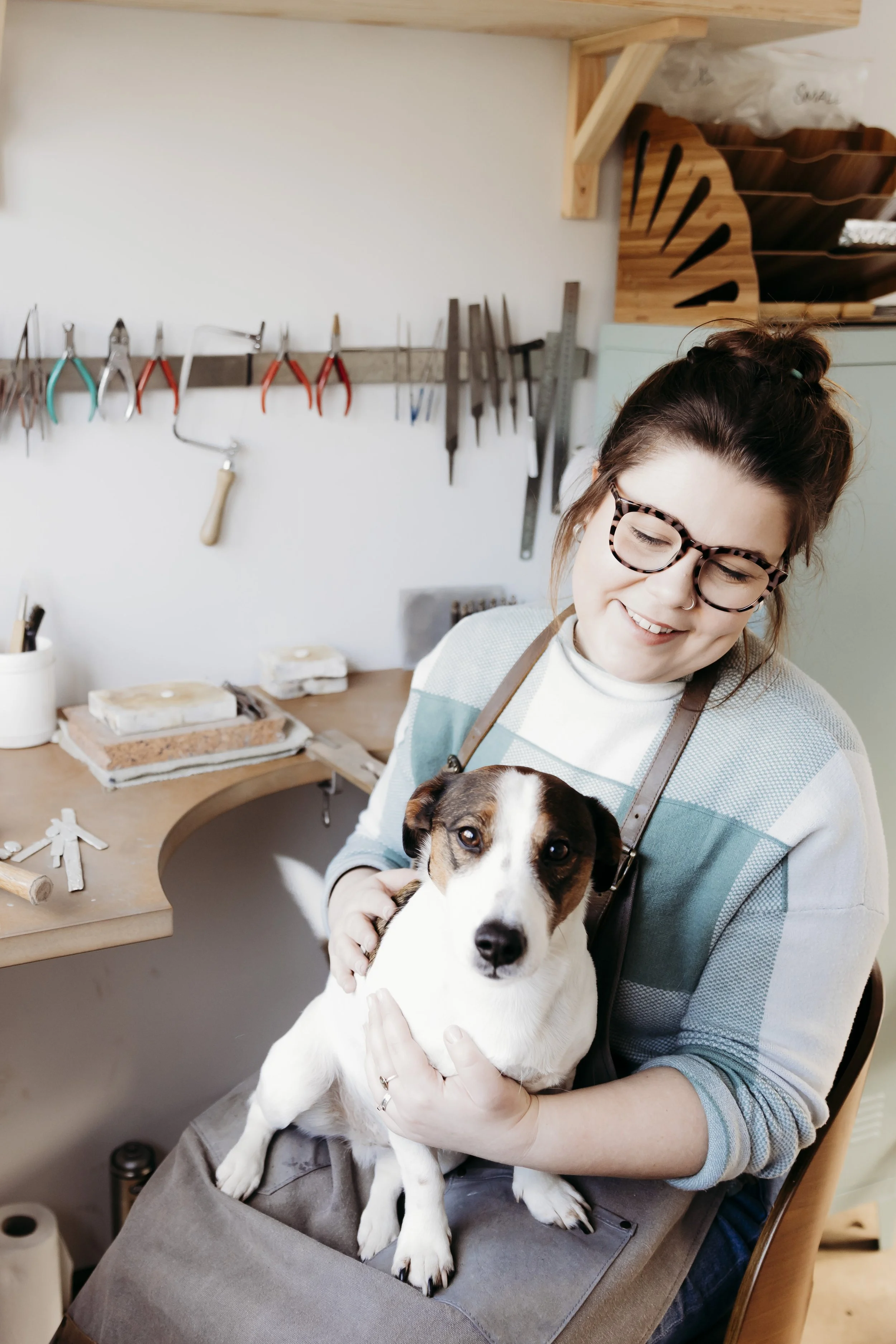 A woman with glasses holding a black and white dog in a grooming salon. The background shows grooming tools hanging on the wall and woodworking supplies on the counter.